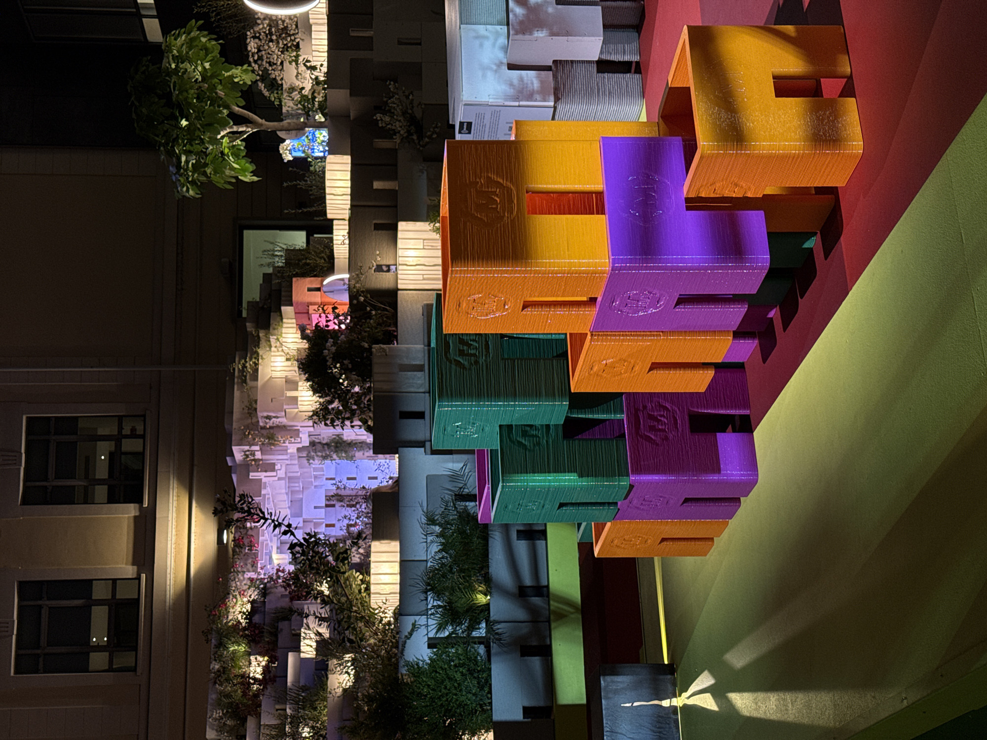 The image captures an illuminated outdoor scene at night, likely an art installation or architectural exhibit in Milan, Italy. The central focus is a towering structure made of large, vibrantly colored, interlocking block-like pieces, predominantly orange, purple, green, and yellow. These blocks appear 3D-printed, showing distinct layer lines, and each features a repeated, embossed logo resembling a stylized "E" or "W" on its surfaces. The structure is set against a backdrop of red and lime green walls, creating strong color contrasts.
In the mid-ground and background, there are architectural elements including buildings with windows, hinting at an urban environment. A prominent feature is a multi-layered wall constructed from numerous rectangular, white or light grey blocks, some of which are illuminated with soft, colorful lights (blue, pink) and others featuring projected patterns or images, such as cloud-like forms. Various trees, bushes, and potted plants are strategically placed throughout the scene, adding natural elements and texture, many also lit by spotlights.
Artificial lighting, including circular overhead lamps and wall-mounted fixtures, casts a warm glow and creates interesting shadows, enhancing the nighttime ambiance. No people are visible in the scene. On one of the white background blocks, an informational sign with black text is partially visible, and the word "Lego" can faintly be discerned, suggesting a connection to the well-known toy company or a similar modular design concept. The overall impression is one of a modern, playful, and visually engaging display.