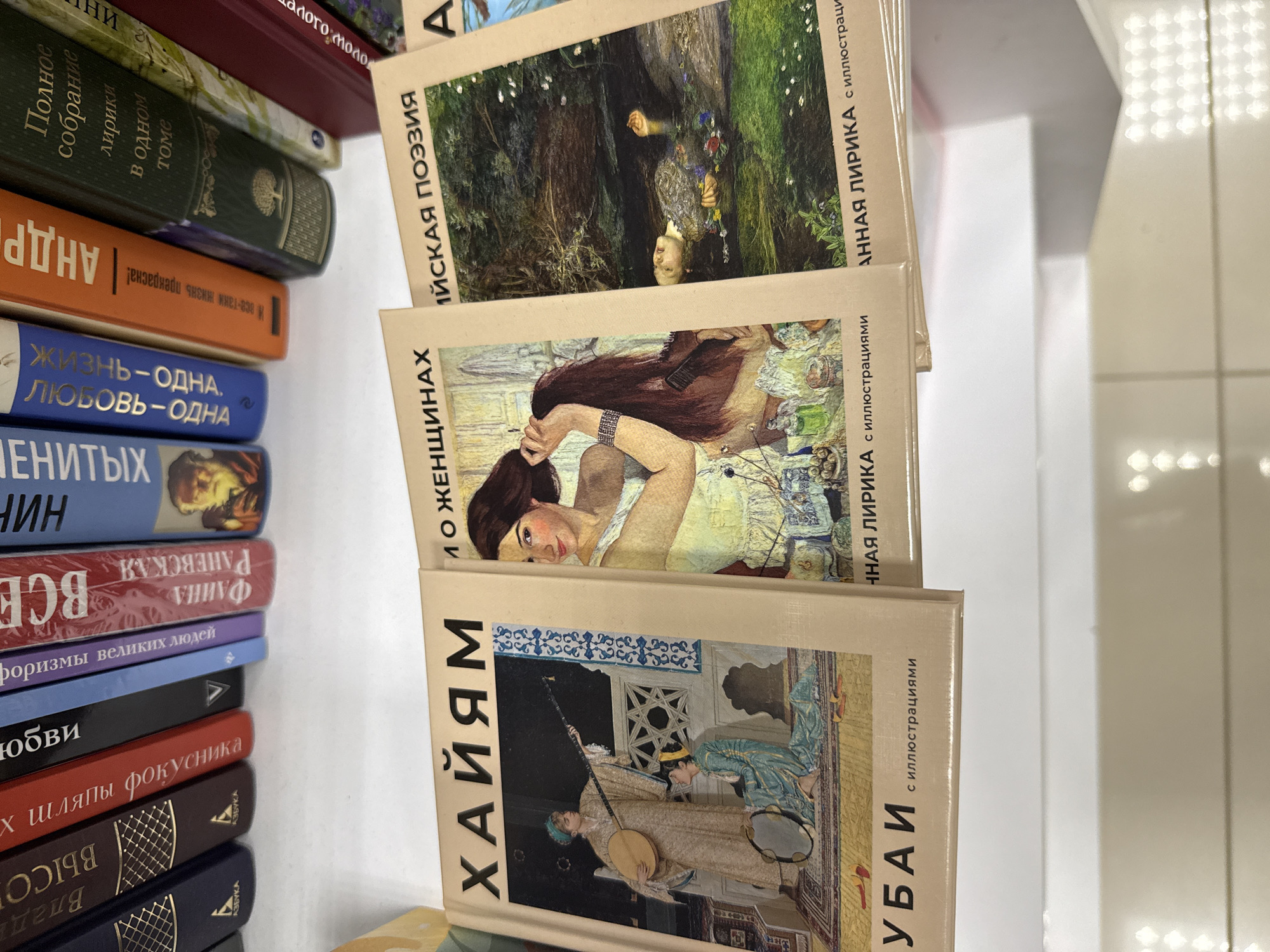 A close-up, high-angle shot shows a shelf with several books in Russian. The books are stacked or leaning against each other, with their spines and covers visible. The books in the foreground have colorful illustrations on their covers. The background consists of blurred shelves and possibly other books. The lighting is bright, creating some glare on the surfaces. There are no people or animals visible in the frame. The scene is likely indoors, possibly in a bookstore or library.