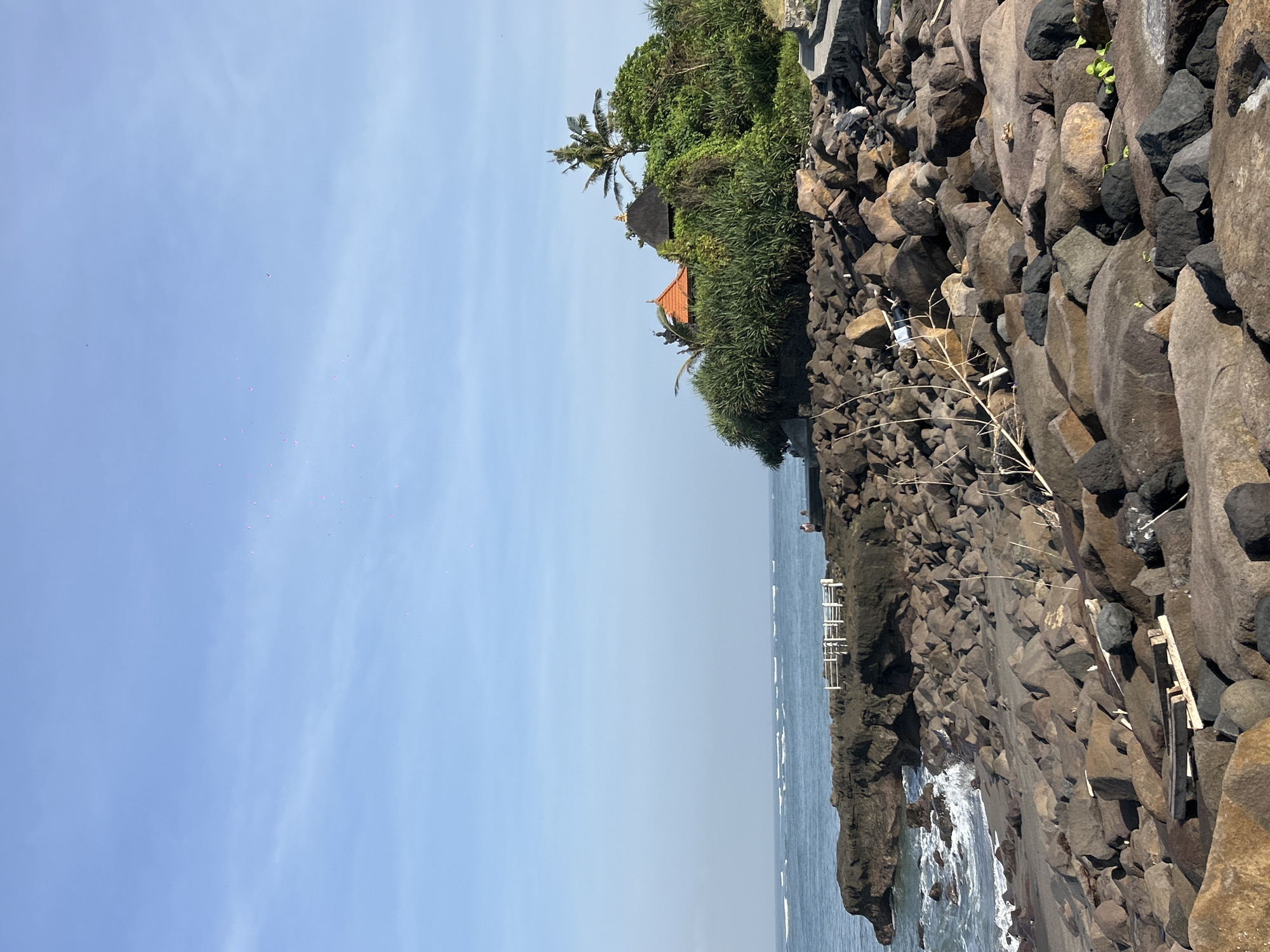 The image shows a coastal scene in Mengwi, Indonesia. The foreground is dominated by large, dark rocks forming a barrier along the shoreline. These rocks transition into a vegetated area with trees, palms, and a building with a red-tiled roof partially visible amongst the greenery. Beyond the rocks, the ocean stretches out to the horizon, meeting a light blue sky with scattered clouds. A white structure, potentially a fence or railing, runs along the edge where the rocks meet the water. A person appears to be standing near the railing.