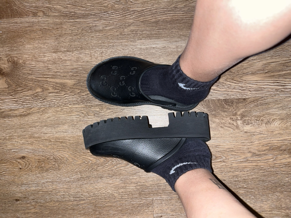 An overhead view depicts two human feet, clad in black socks and shoes, positioned on a light brown wooden floor. The scene is located in Middelburg, Netherlands.
The subject's legs are visible from the lower calf downward, exhibiting light-toned skin. The left foot wears a black ankle sock with a white Nike swoosh logo visible on the outer side, and a faint, dark-inked tattoo is present on the outer aspect of the lower left leg, above the ankle. This foot is partially inserted into a black, open-backed shoe, identified as a clog or mule with a thick, ridged black platform sole. The upper of the left shoe appears to be made of a textured, possibly grained material.
The right foot also wears a black ankle sock with a white Nike swoosh logo on the outer side. This foot is fully inserted into a black, slip-on shoe. The upper surface of the right shoe is embossed with multiple "GG" logos, indicating a Gucci brand item.
The flooring consists of horizontal wooden planks, displaying natural grain patterns and knots. No other objects or environmental details are discernible. The feet are positioned slightly apart, angled to present the footwear.
