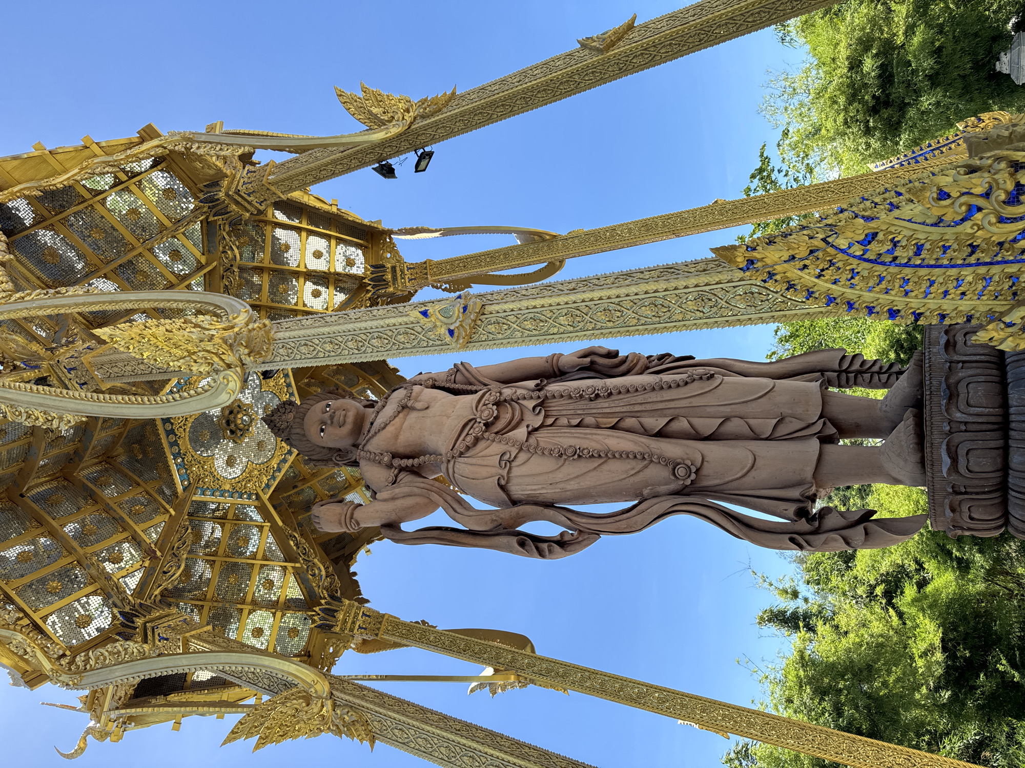 A statue of a standing deity stands under an ornate golden canopy in Bang Pu Subdistrict Municipality, Thailand. The canopy is supported by four gilded pillars, decorated with intricate designs and blue accents, reaching towards a clear blue sky. Lush green trees are visible in the background. The deity statue, seemingly made of a light brown material, features detailed robes, necklaces, and facial features.