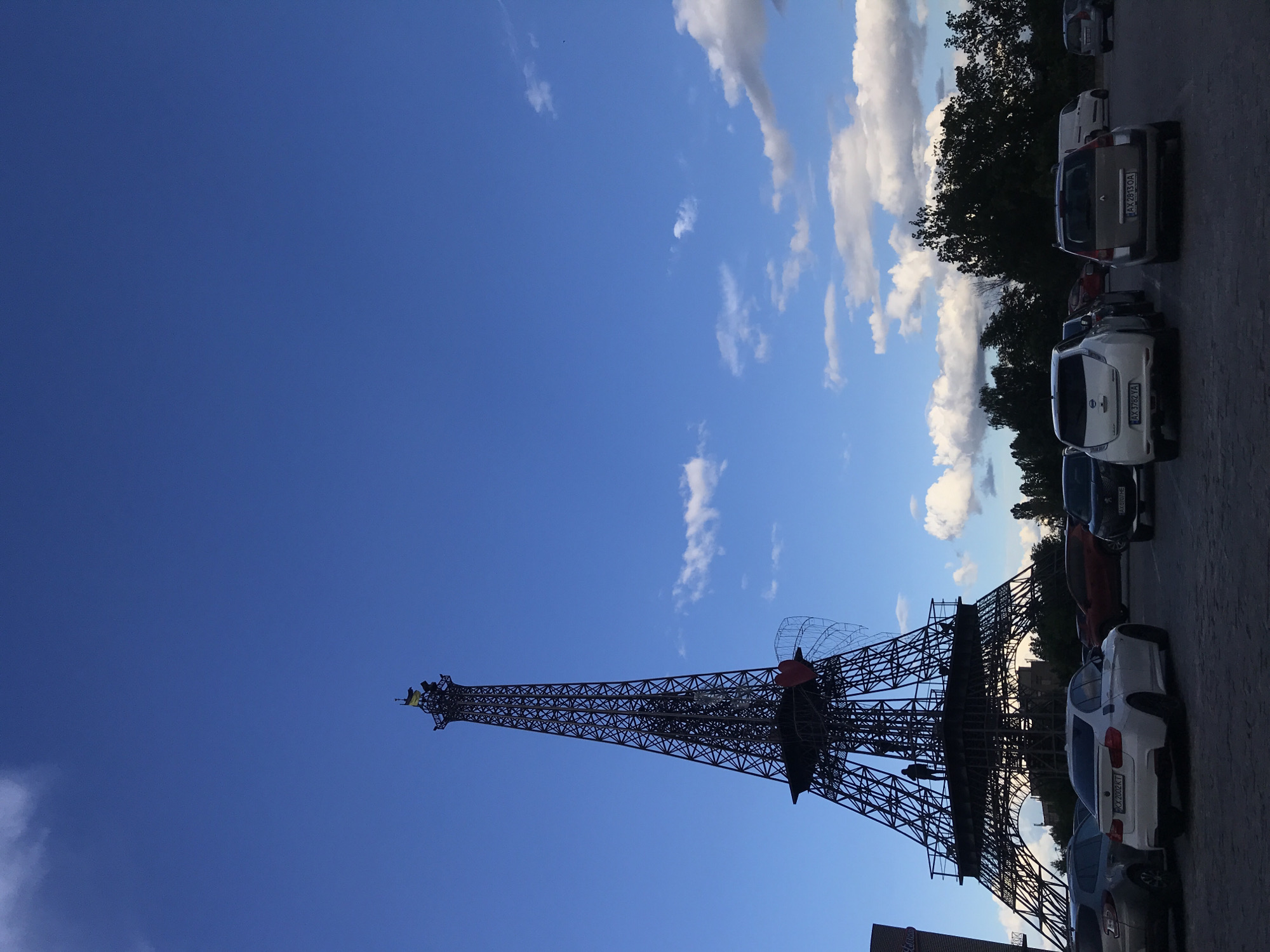 The image presents a low-angle view of a replica of the Eiffel Tower, positioned next to a row of parked cars on a street in Kharkiv, Ukraine. The sky is predominantly blue with scattered white clouds. The Eiffel Tower replica is made of black metal and appears to be located in a parking lot or public space. Several cars are parked to the right of the tower, and trees are visible along the upper-right edge of the photo.
