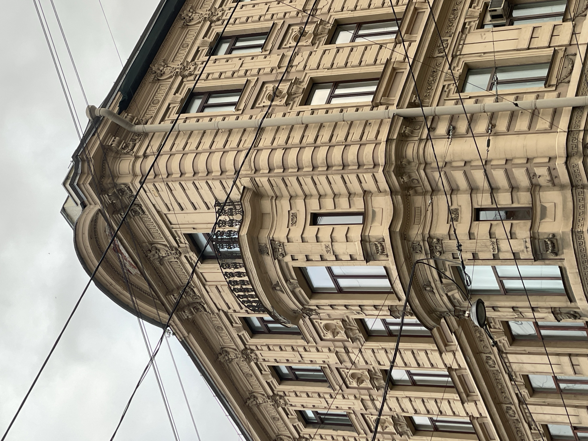 The image shows the exterior of a multi-story building in Saint Petersburg, Russia, likely in the historical center. The building is ornate, featuring decorative facades, windows, balconies, and molding. The sky is overcast. Power lines and drain pipes are visible against the backdrop of the building. The building appears to be constructed with sandstone or a similar light-colored material.