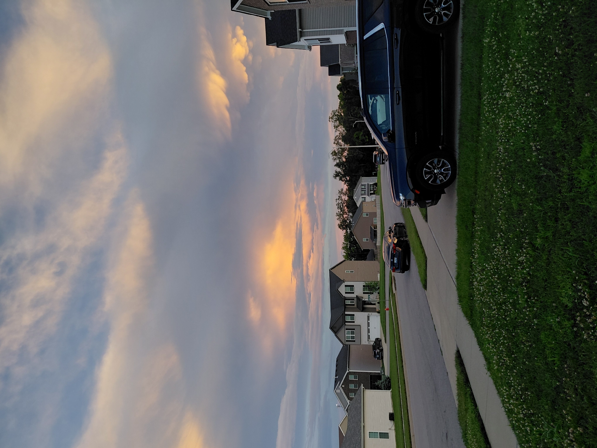 This is a photograph taken at dusk in Nicholasville, Kentucky, USA. The sky is filled with dramatic clouds, illuminated by the setting sun with hues of orange, yellow, and gray. In the foreground, a dark-colored SUV is parked on the right side of the frame, partially obscuring a well-maintained lawn with white clover. A sidewalk runs along the edge of the lawn, leading to a street. Across the street, several suburban houses are visible, with a dark sedan parked in the driveway of one of them. Trees line the background, silhouetted against the vibrant sky. The overall scene depicts a peaceful residential street at the end of the day.