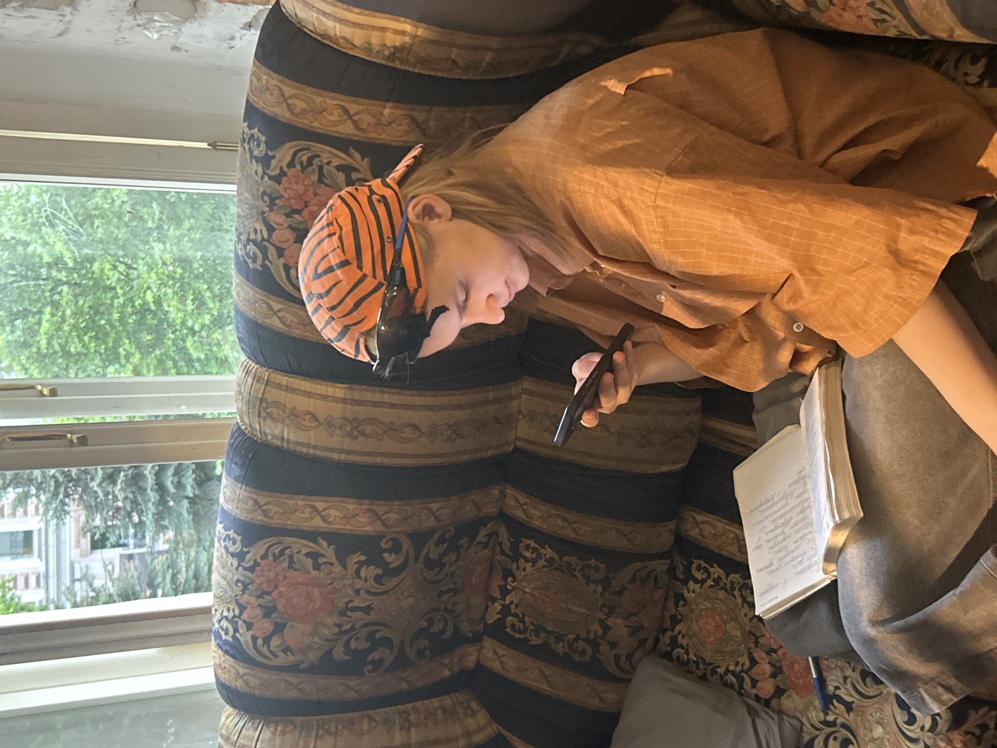 A person wearing an orange and black striped hat is reclining on a floral patterned couch. They are wearing an orange button-up shirt and grey sweatpants. The person is holding a black smartphone in their left hand. On their legs rests an open book. A window is visible in the background, showing trees and buildings outside. The scene appears to be indoors. The location is Unknown, Unknown.