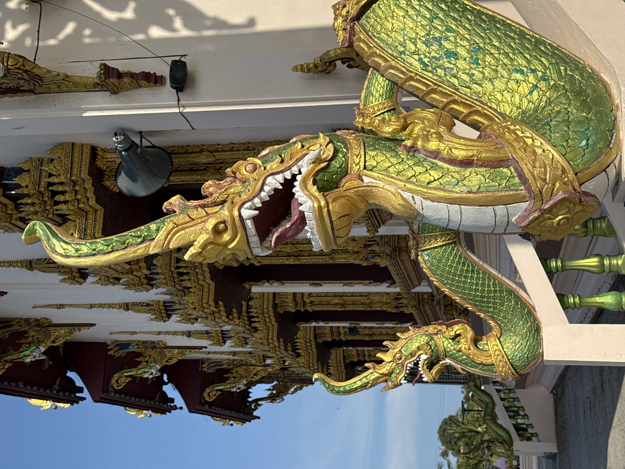 The image shows a close-up view of a building with ornate golden decorations and statues of mythical creatures, likely a temple or shrine in Ko Samui District, Thailand. The foreground features a large, green and gold serpent-like statue with its mouth open, revealing sharp white teeth and a red tongue. This statue appears to be a decorative railing or guardian figure. Behind it, another, smaller serpent statue is visible, also with its head raised.
The building itself is white with prominent gold embellishments, including intricate carvings and tiered roofing. The architectural style suggests a Southeast Asian influence, consistent with temples found in Thailand. A black loudspeaker is mounted on the wall of the building, to the right of the larger serpent statue. The sky is a clear, bright blue, indicating daytime. The overall scene conveys a sense of cultural and religious significance, with the statues serving as decorative and protective elements. The ground in front of the building is paved, and to the left, there are glimpses of trees and foliage, suggesting an outdoor setting.