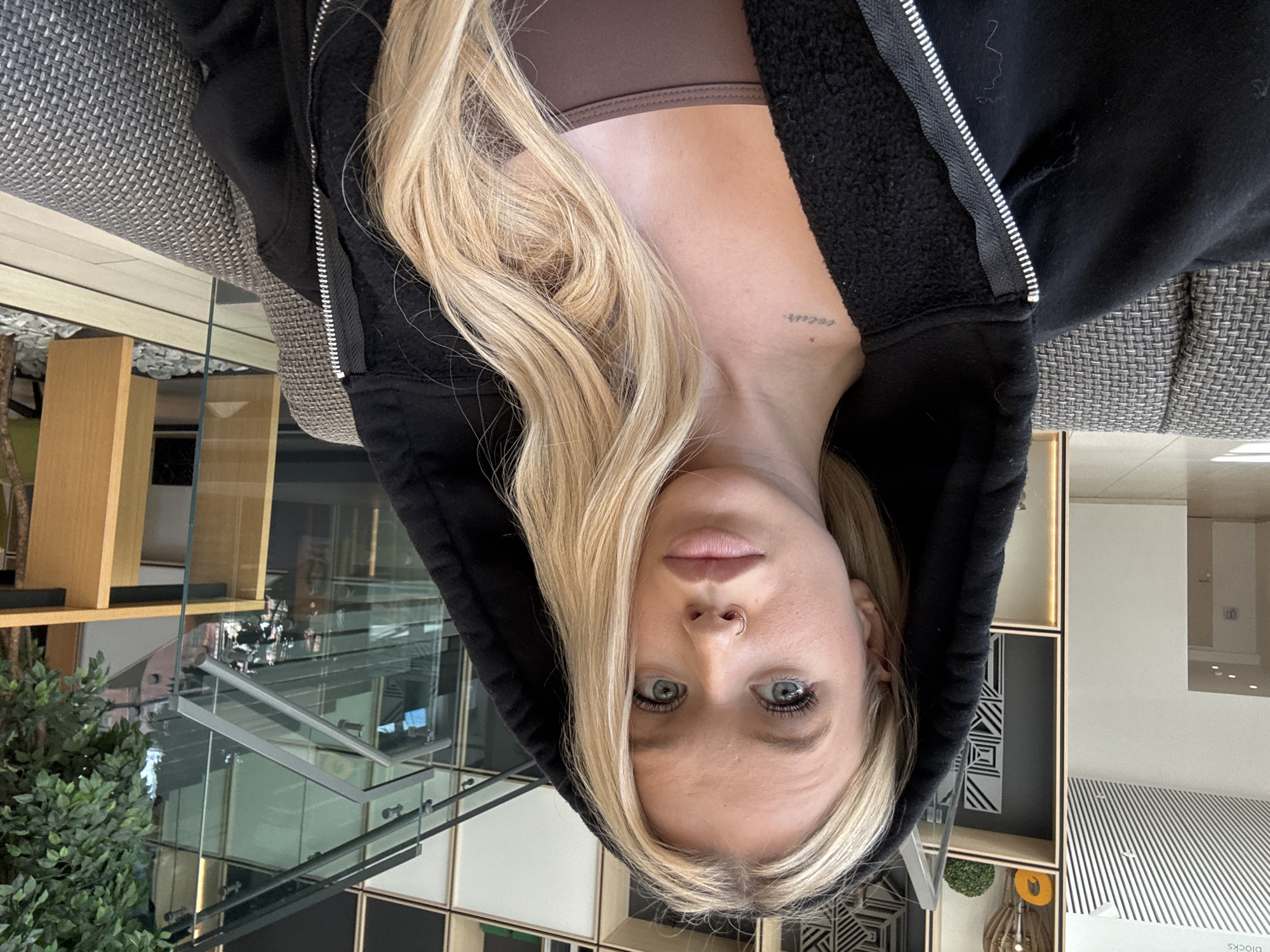 A young woman with blonde hair and blue eyes, wearing a black hooded jacket and a brown top, is shown in a close-up, upside-down portrait. She has a nose ring and a small tattoo on her neck. The background suggests an indoor setting with modern architecture, including glass railings, wooden shelves, and patterned wall decor. The lighting appears to be artificial, indicating it is likely daytime or evening indoors. The woman's expression is neutral, with her gaze directed towards the camera. The overall scene conveys a casual and contemporary atmosphere. The image is a selfie, taken from a low angle. There is no visible text in the image. The location context is Saint Petersburg, Russia.