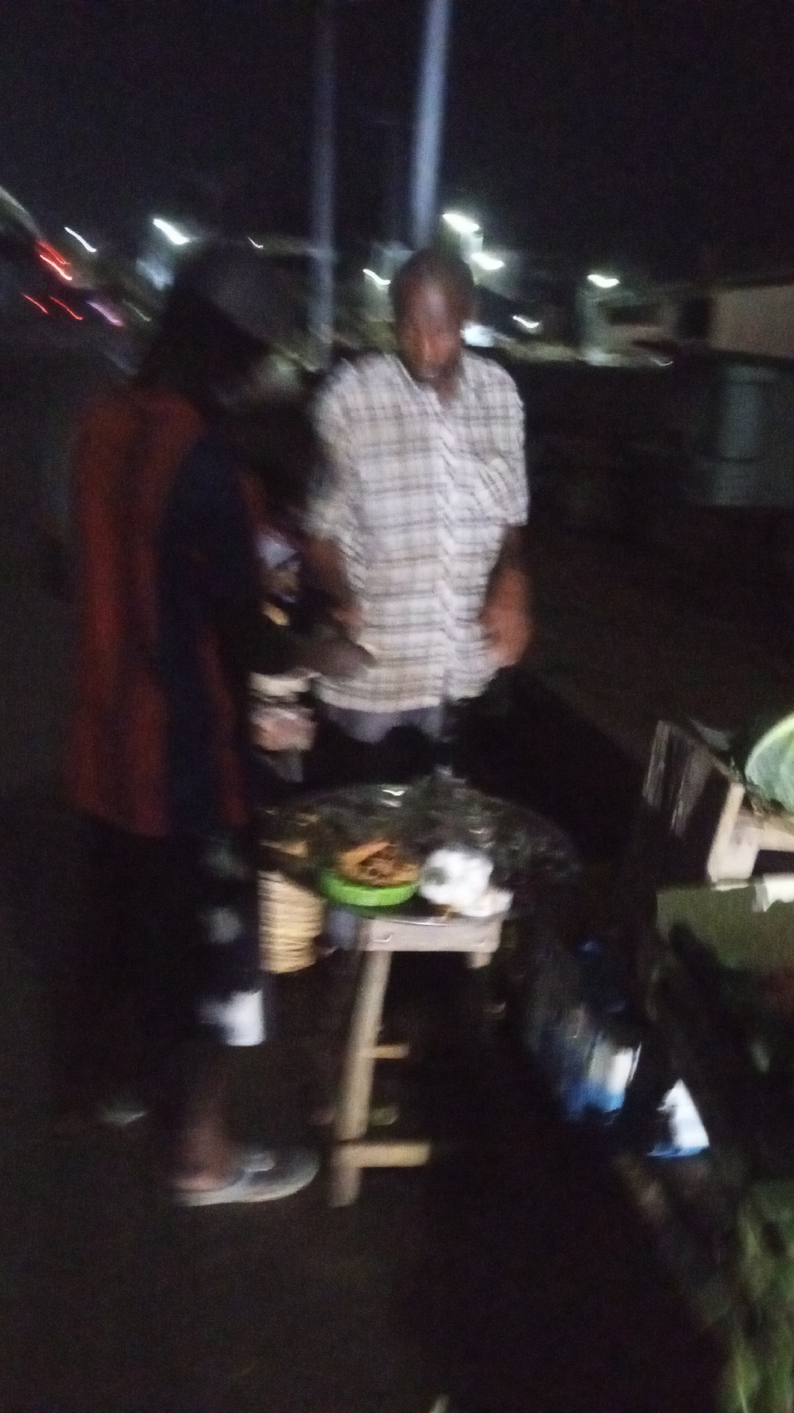 The image depicts an outdoor, low-light scene at night, likely in a street or market in Lokoja, Nigeria. Two individuals are visible, blurred due to the dim conditions. On the left, a person is seen from behind, wearing a dark top, lighter shorts, and flip-flops. Centrally, another person, potentially a vendor, wears a light-colored plaid shirt and appears to have a beard; they are focused on items laid out on a small, light-colored wooden stool or table.
The stool serves as a makeshift display, holding a bright green bowl containing what appears to be fried food items, possibly plantains or similar snacks. Nearby on the stool are several white-wrapped packages, suggesting additional food items ready for sale. A woven basket rests on the ground to the left of the stool. The background is dark, punctuated by blurred streaks of light from what might be streetlights or passing vehicles, characteristic of an active nighttime street. The scene captures a candid moment of a street vendor attending to their goods, possibly interacting with a customer. No visible text is present in the image.
