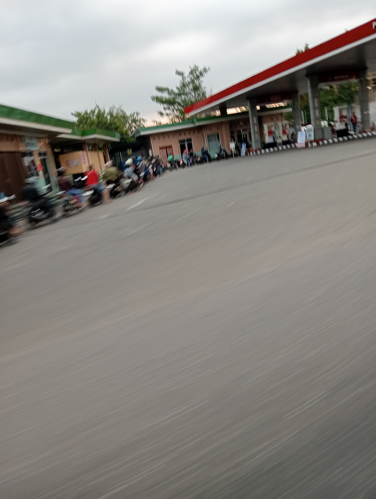 The image depicts a blurry scene at a gas station in Danurejo, Indonesia. The gas station has a red and white canopy supported by columns. Several motorcycles are parked in a line, with people sitting on them, suggesting a queue. Buildings with green roofs are visible on the left side of the image, and trees can be seen behind the gas station. The sky is overcast. The overall impression is that of a busy, but somewhat chaotic, gathering at the gas station, likely due to fuel shortages or a high demand for gasoline.