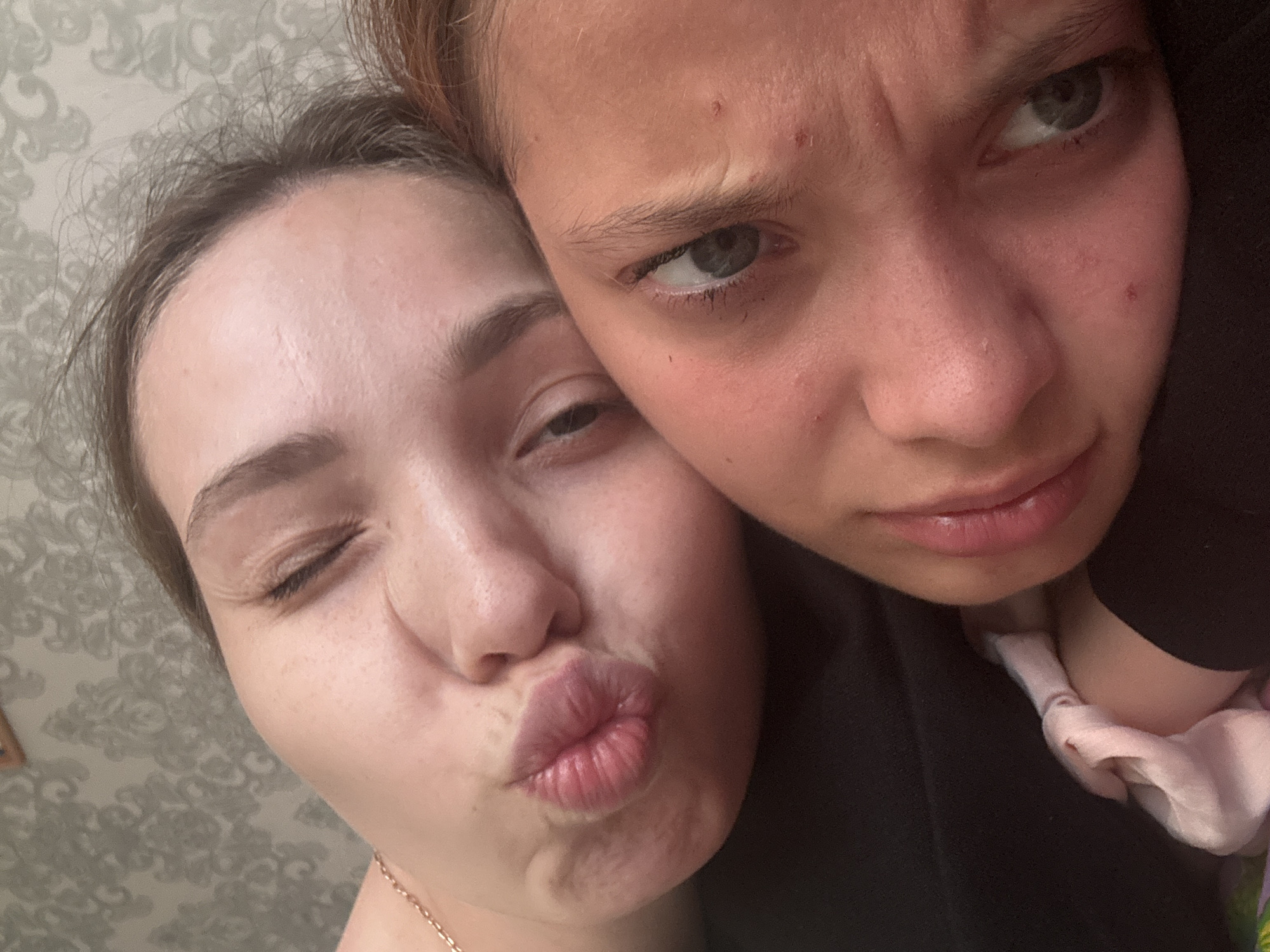 This is a close-up, tilted, selfie-style photograph featuring two young women in an indoor setting. The woman in the foreground on the right has her head tilted back, with her eye looking slightly upward and to the left. She has light skin with some blemishes and dark eyelashes. The woman in the background on the left is also tilted, with her face angled towards the camera. She is winking, with one eye closed and the other open, and her lips are puckered in a kiss pose. She has a delicate gold chain visible around her neck. The background appears to be a wall with patterned wallpaper. The lighting is soft, suggesting an indoor environment. There is no indication of the city of Kemul, Russia, or any other specific location.