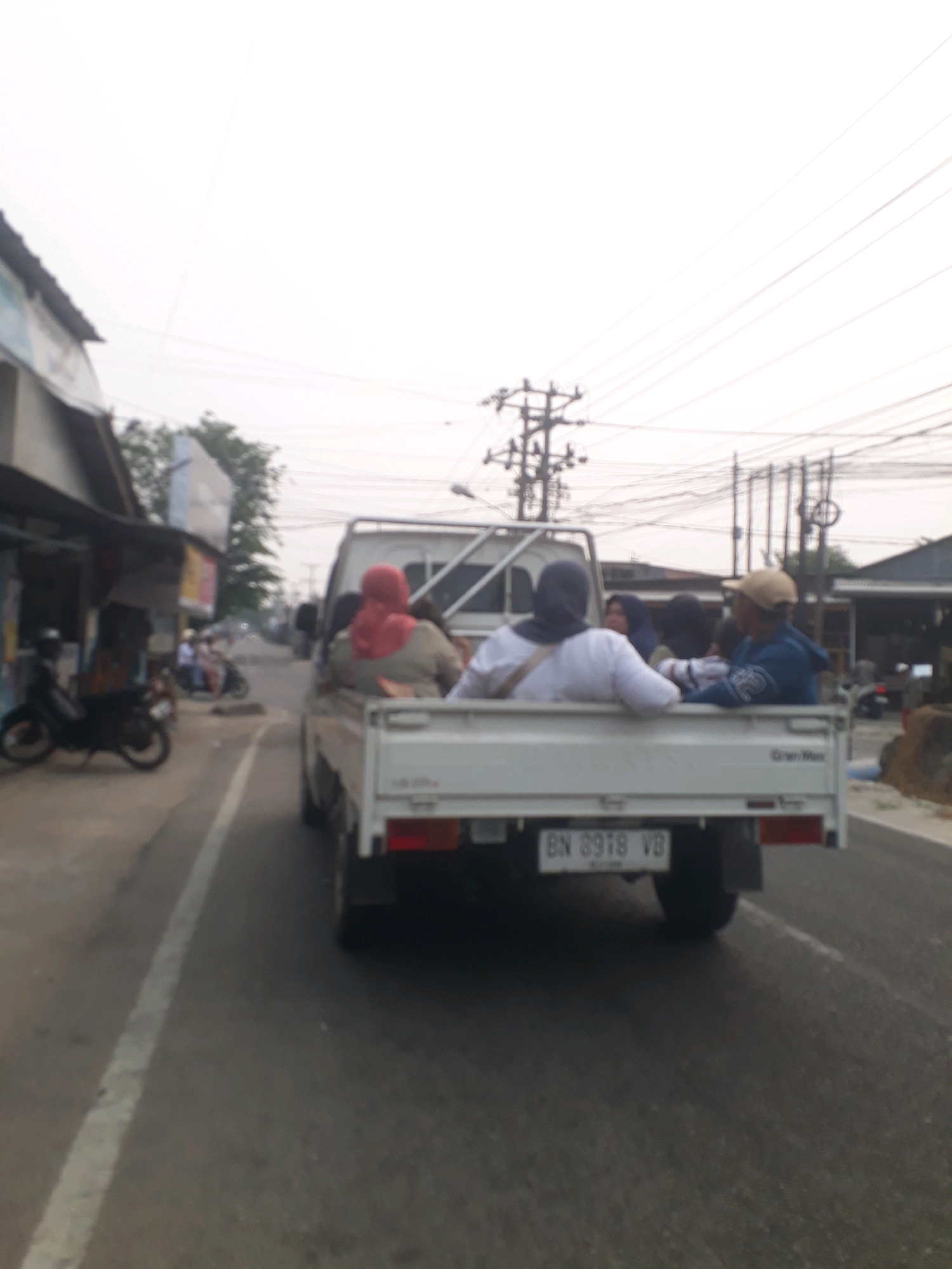 In Pangkalpinang, Indonesia, a white pickup truck with passengers in the bed travels on a paved road. The truck's license plate reads "BN 6918 VB." The truck is loaded with at least four people, two women in hijabs, one wearing a red hijab and the other a dark blue hijab, and two men. One man wears a beige cap and a blue hooded jacket, and the other is partially obscured. The truck appears to be on a street with shops on the left side and utility poles and wires overhead on the right. The sky is overcast, and the overall lighting suggests it is daytime. The image is somewhat blurry, indicating movement.