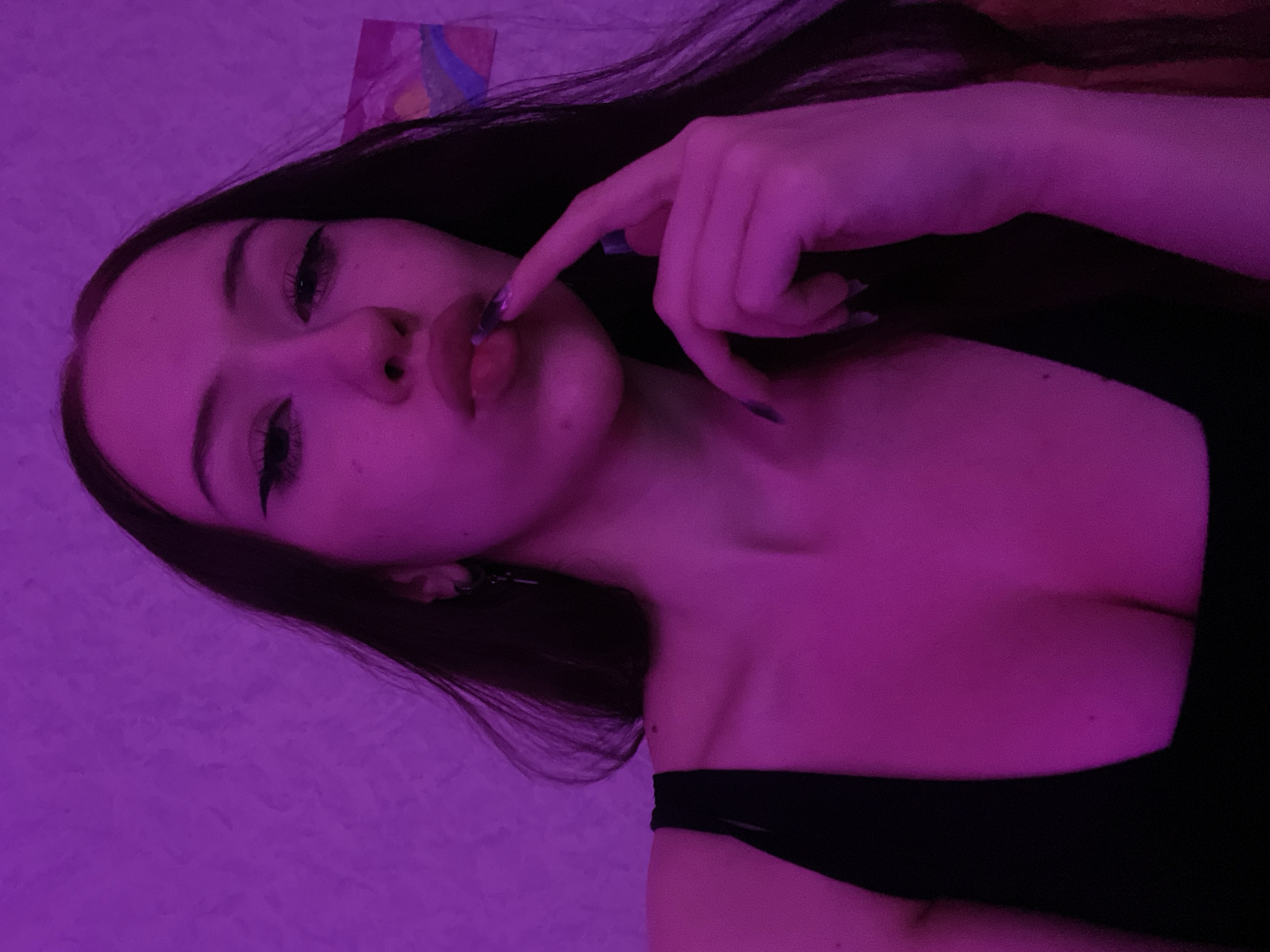 The image shows a close-up, waist-up shot of a young woman with long, dark hair, taken indoors with purple lighting. She is wearing a black tank top that reveals her décolletage. Her right hand is brought to her lips, with her index finger touching her lower lip. She has dark eyeliner and bold eyebrows, and her lips are slightly parted. Her nails are long and dark. The background is a textured wall illuminated by a purple light source, with a piece of artwork visible on the right. There is no indication of Votkinsk, Russia, or any specific city in the image.