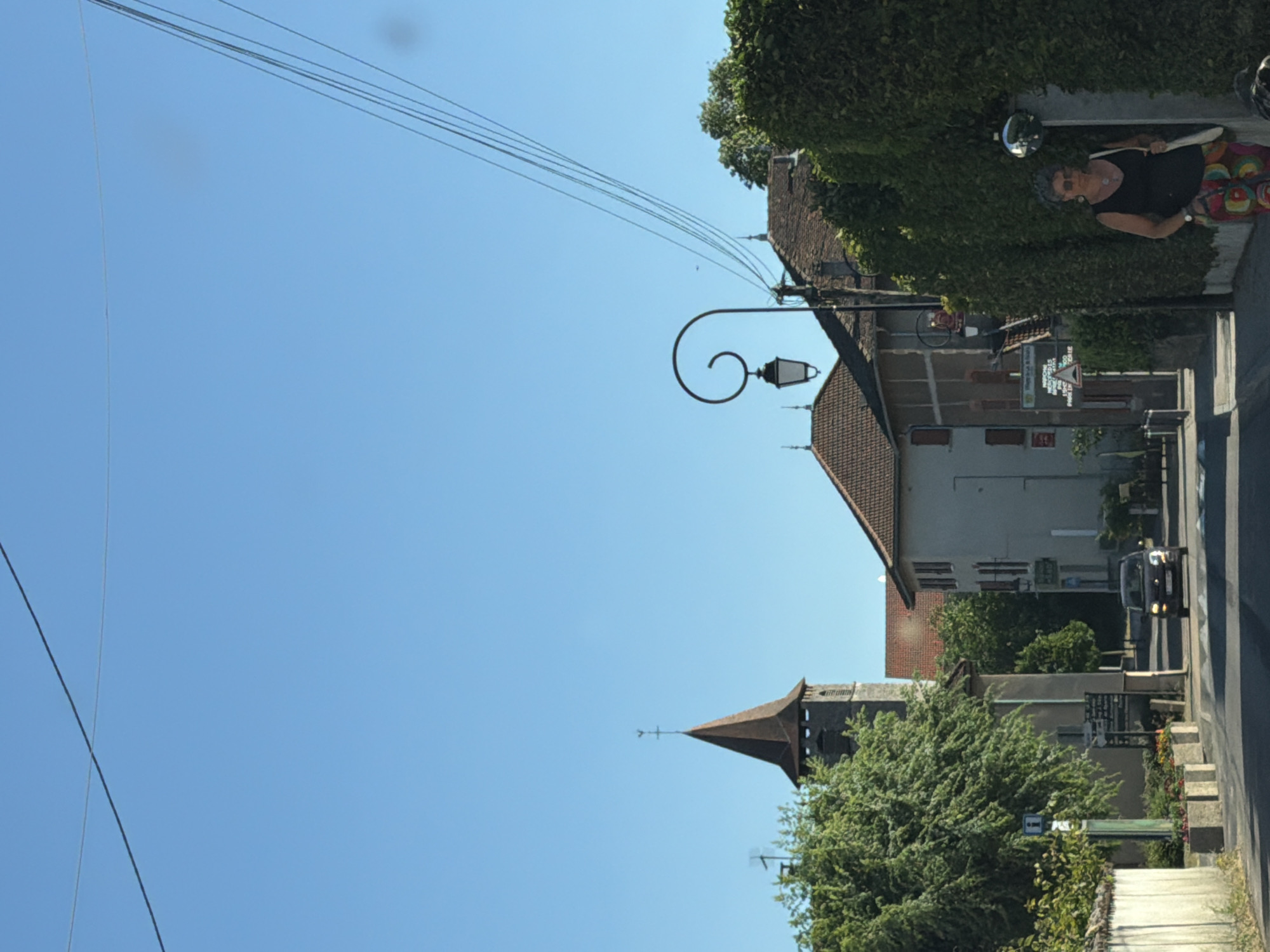 A street scene in the city of Villages du Lac de Paladru, France, shows a vehicle driving down a paved road lined with buildings and lush green foliage under a clear blue sky. A church spire with a weather vane is visible to the left, while buildings with red-tiled roofs and a street lamp adorn the right side of the road. A woman in a black top and colorful patterned pants walks on the right side of the road. Signs with French text are posted along the street.