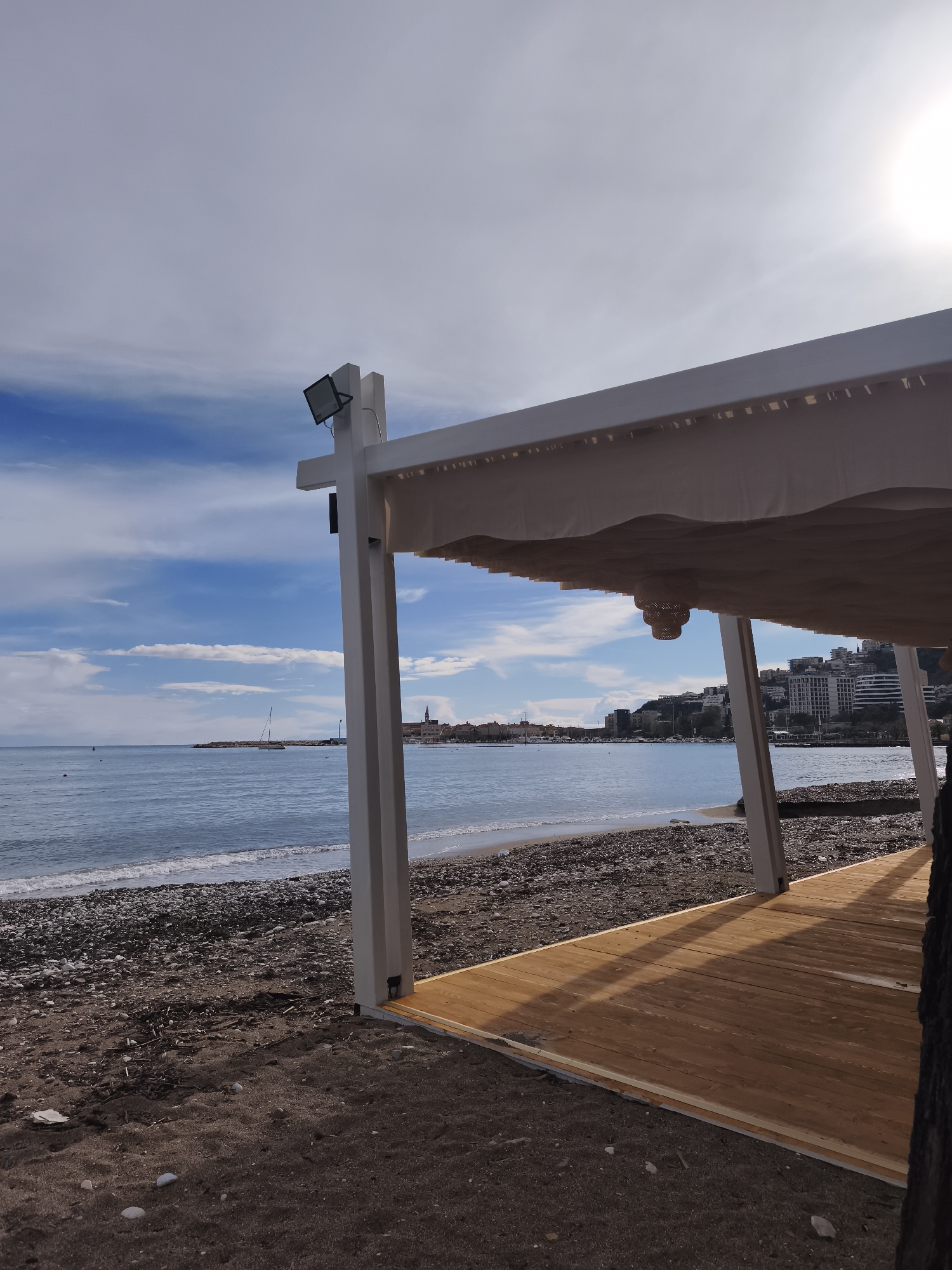 The image depicts a coastal scene in Budva, Montenegro, observed from the perspective of a beach.
In the immediate foreground on the right, a white, modern pergola structure with a light-colored retractable fabric canopy is partially visible, extending from the bottom right corner towards the upper right. A black rectangular spotlight is mounted on the upper left pillar of this structure. Directly below the canopy, a woven, beige, bell-shaped light fixture hangs. A light brown wooden deck, composed of parallel planks, is partially visible beneath the canopy on the lower right. The ground in front of and around the structure is dark sandy earth mixed with small, dark pebbles and some light-colored debris.
The midground features a coastline with waves gently breaking on a beach primarily composed of small, dark pebbles transitioning to dark sand closer to the foreground. The sea extends horizontally across the frame, exhibiting a light blue hue. In the water to the left of the center, a white sailing yacht with a single mast is moored. Further left and closer to the shore, a small orange buoy is visible, and two indistinct dark figures are in the water near it, appearing to be swimming or wading. A rocky breakwater or landmass extends from the left coastline into the sea.
In the background, across the bay, the historic architecture of Budva Old Town (Stari Grad) is visible on the left, distinguished by its traditional buildings and a prominent church spire. To the right, modern multi-story buildings and hotels are situated on a sloping hill. The sky overhead is a mix of clear blue and scattered, wispy white clouds. A bright sun is visible in the upper right corner, casting a glare.