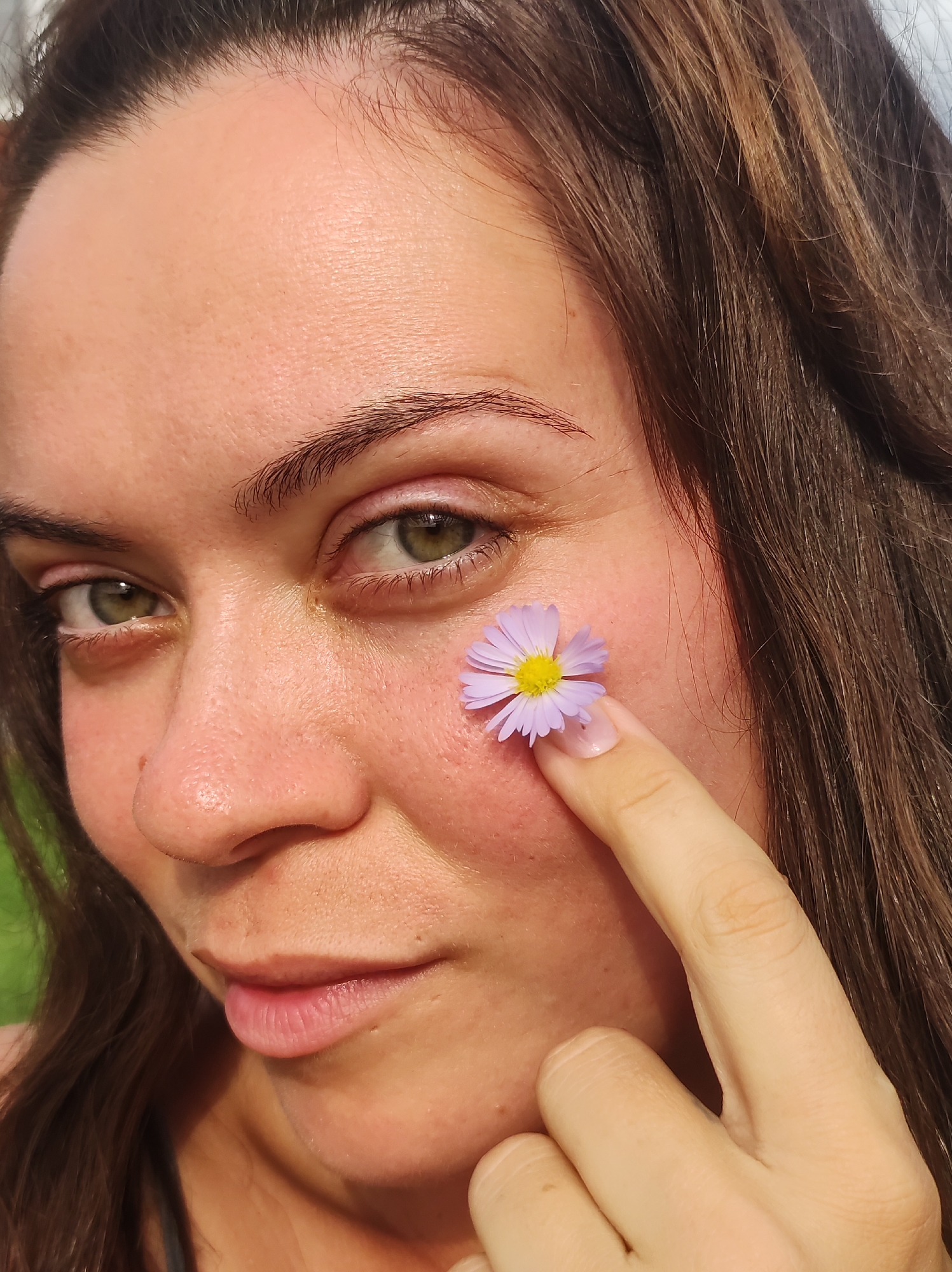 This is a close-up, waist-up, natural light, outdoor portrait of a woman with brown hair and green eyes. She is holding a small purple and yellow flower against her cheek with her right index finger. Her skin is fair with some visible pores and freckles. The background is blurred green, suggesting an outdoor, natural setting, possibly in the city of Fokino, Russia, given the context provided. The overall mood is serene and natural.