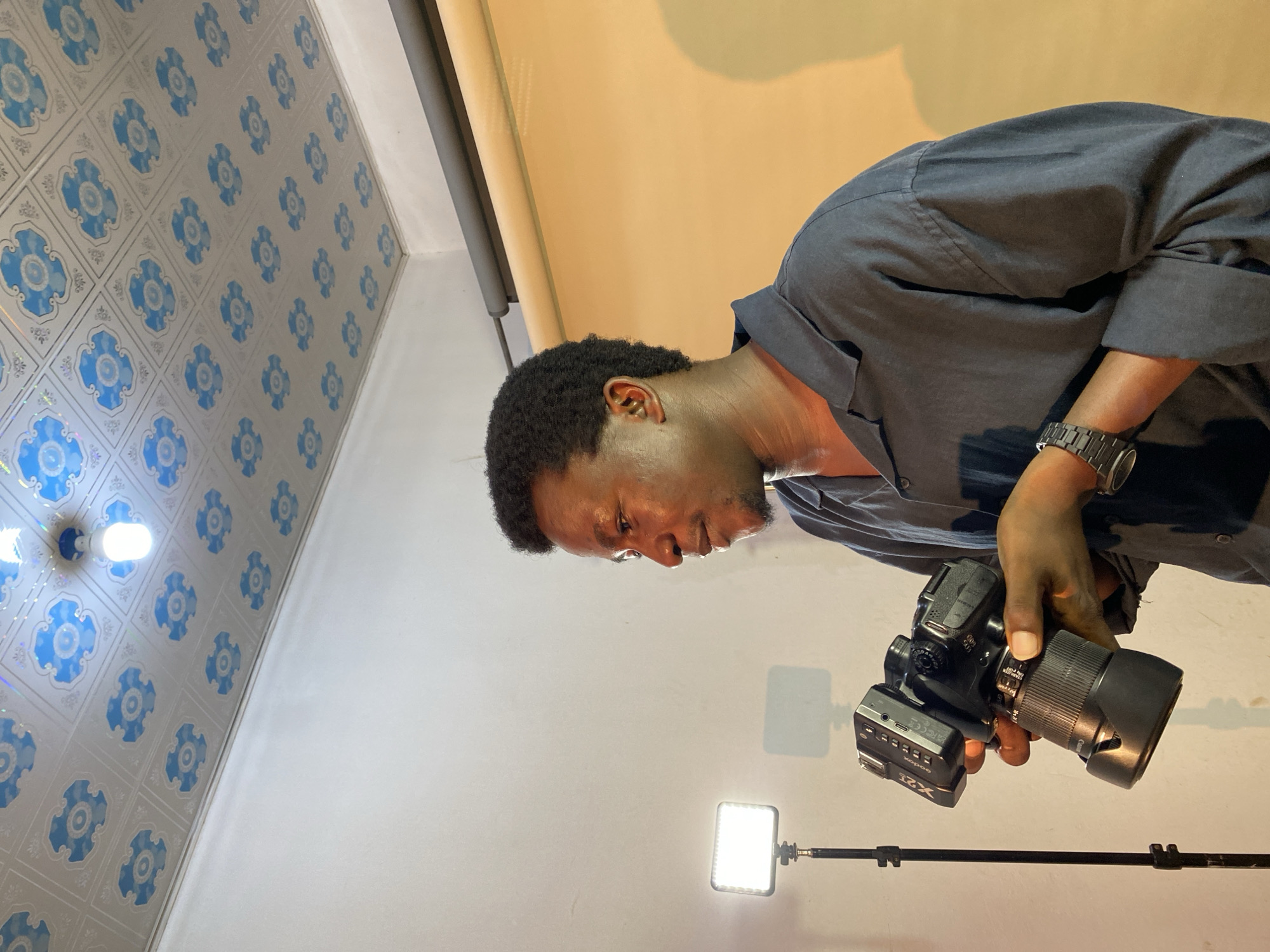 In Gura Top, Nigeria, a man stands in a room, holding a camera. He is wearing a dark collared shirt and a watch. In the background, a light stand with a white LED panel is visible, casting a shadow on the wall. The room has a white wall and a ceiling with a decorative pattern. The man is positioned in front of a tan backdrop.
