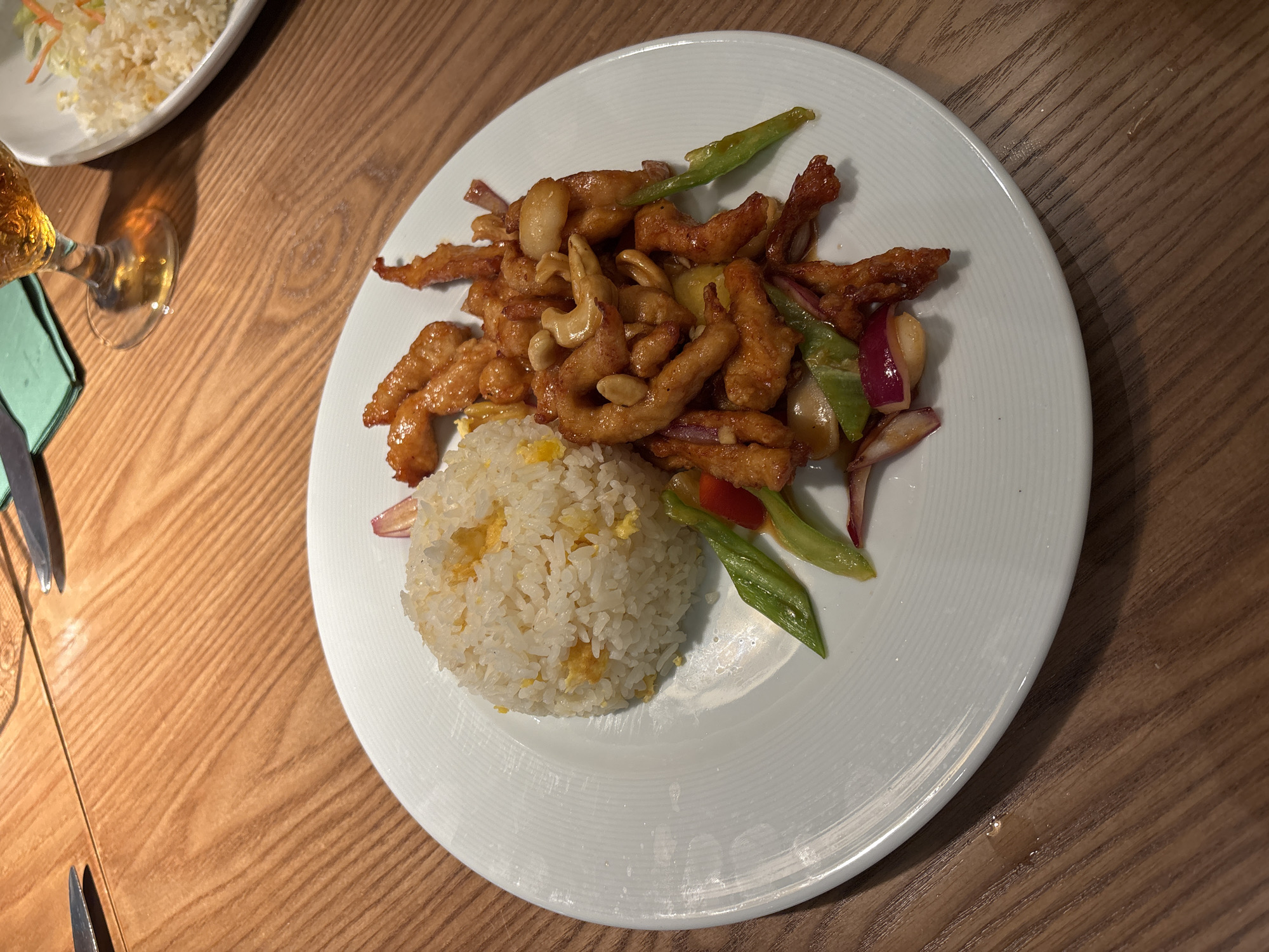 A white plate of food sits on a wooden table. The plate contains fried chicken, cashews, green peppers, and red onions, served with a mound of white rice. The food is presented in a dish that is predominantly viewed from a top-down perspective. The background shows a glass of amber liquid, a green napkin, and cutlery. The scene is lit by ambient lighting. The food appears to be a meal served at a restaurant. The cuisine is likely Asian, given the components of the dish. The table is made of wood, with visible grain patterns and a smooth finish. The cutlery is silver, and the glass is clear.