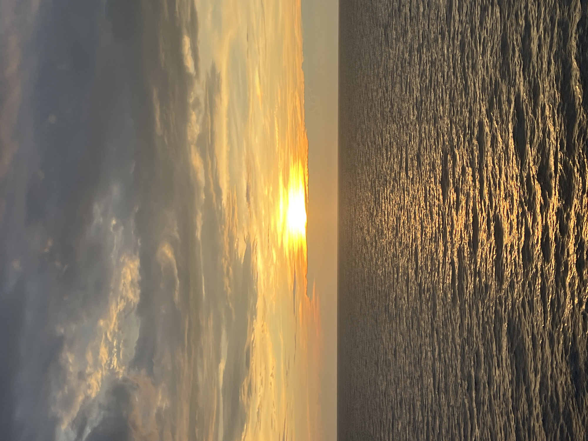 A wide-angle shot captures the vast expanse of the ocean under a dramatic sunset. The horizon line is clearly defined, separating the deep blue-grey water from the sky. The ocean's surface is textured with small ripples, reflecting the warm golden light of the setting sun. The sky is a canvas of color, with darker grey clouds dominating the upper portion, transitioning to vibrant orange and yellow hues as they approach the horizon where the sun is partially obscured by a thin band of clouds. The sun itself is a brilliant, bright point of light, casting a shimmering golden pathway across the water. The scene is devoid of any human presence or artificial structures, suggesting a remote or natural setting.