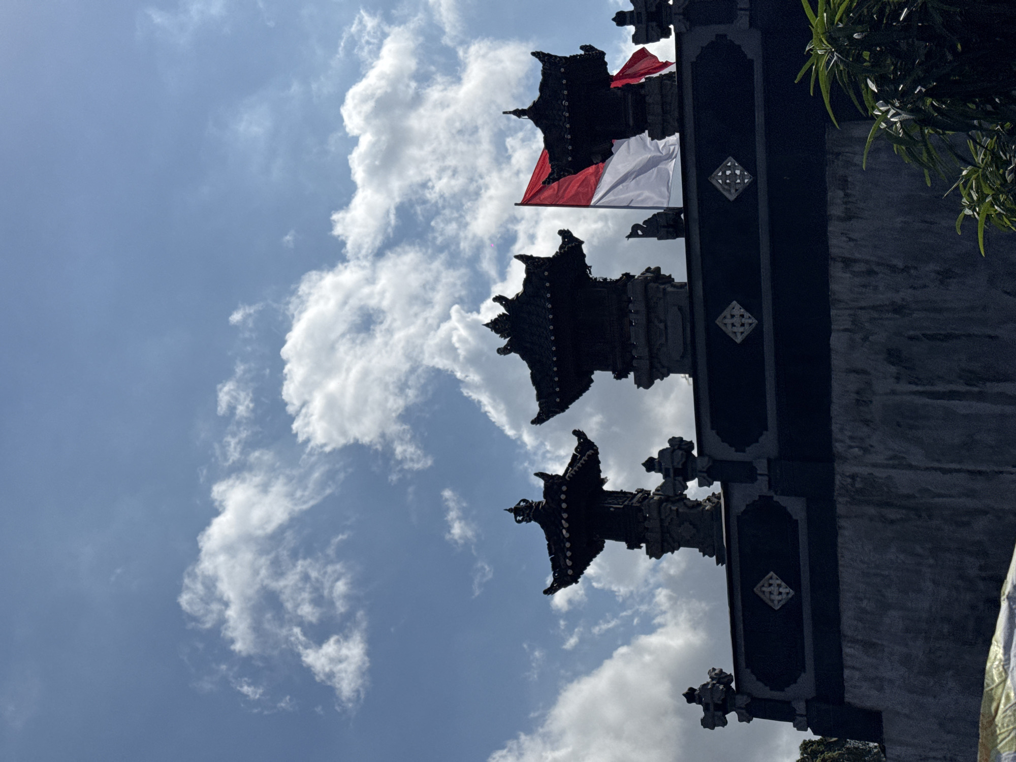 The image shows a bright blue sky with scattered white clouds. In the foreground, silhouetted against the sky, is the upper portion of a dark-colored building, likely a temple or traditional structure, with ornate tiered roofs. An Indonesian flag, red and white, is prominently displayed, fluttering in the wind on a flagpole to the right of the central roof structure. The building's facade features decorative diamond shapes. To the right and lower portion of the image, lush green foliage is visible. The scene appears to be outdoors on a sunny day, with the architectural style suggesting a location in Indonesia, possibly in Denpasar given the presence of the Indonesian flag and traditional architecture.