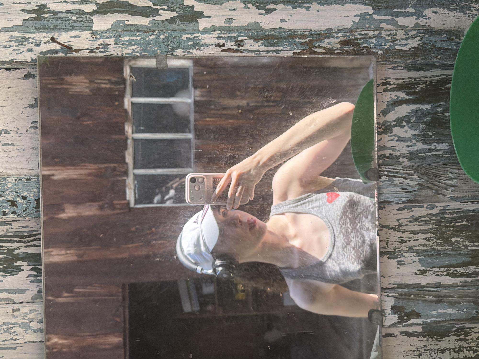 A woman takes a mirror selfie outdoors, wearing a grey tank top with a red heart design and a white baseball cap, with headphones on. She is holding a pink-cased smartphone in her right hand. The reflection shows a rustic wooden building with a barred window behind her. The exterior of the mirror frame is weathered and peeling, with a nail visible on the right side. The scene appears to be in a rural or outdoor setting, possibly a shed or barn, but no specific location in Zavodoukovsk, Russia can be determined from the visual information.