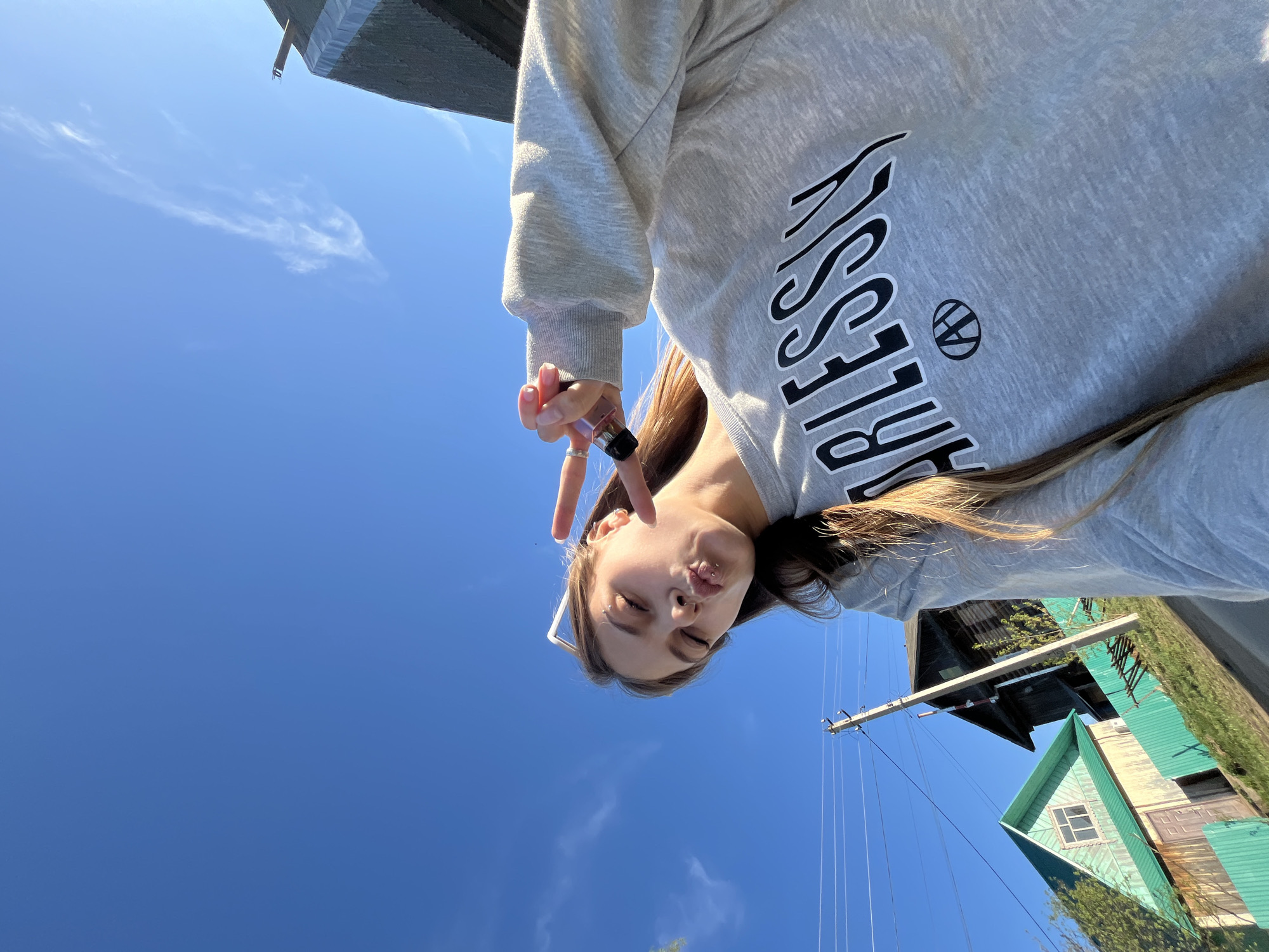 The image shows a young woman taking a selfie from a low angle. She's wearing a gray sweatshirt with "BLESSING" printed on it and making a peace sign. She has light brown hair and is wearing glasses. The background includes a clear blue sky with some wispy clouds, a green building with a window, and a utility pole with wires. The photo appears to be taken outdoors in a residential area. The photo appears to be taken near Фоки, Russia.