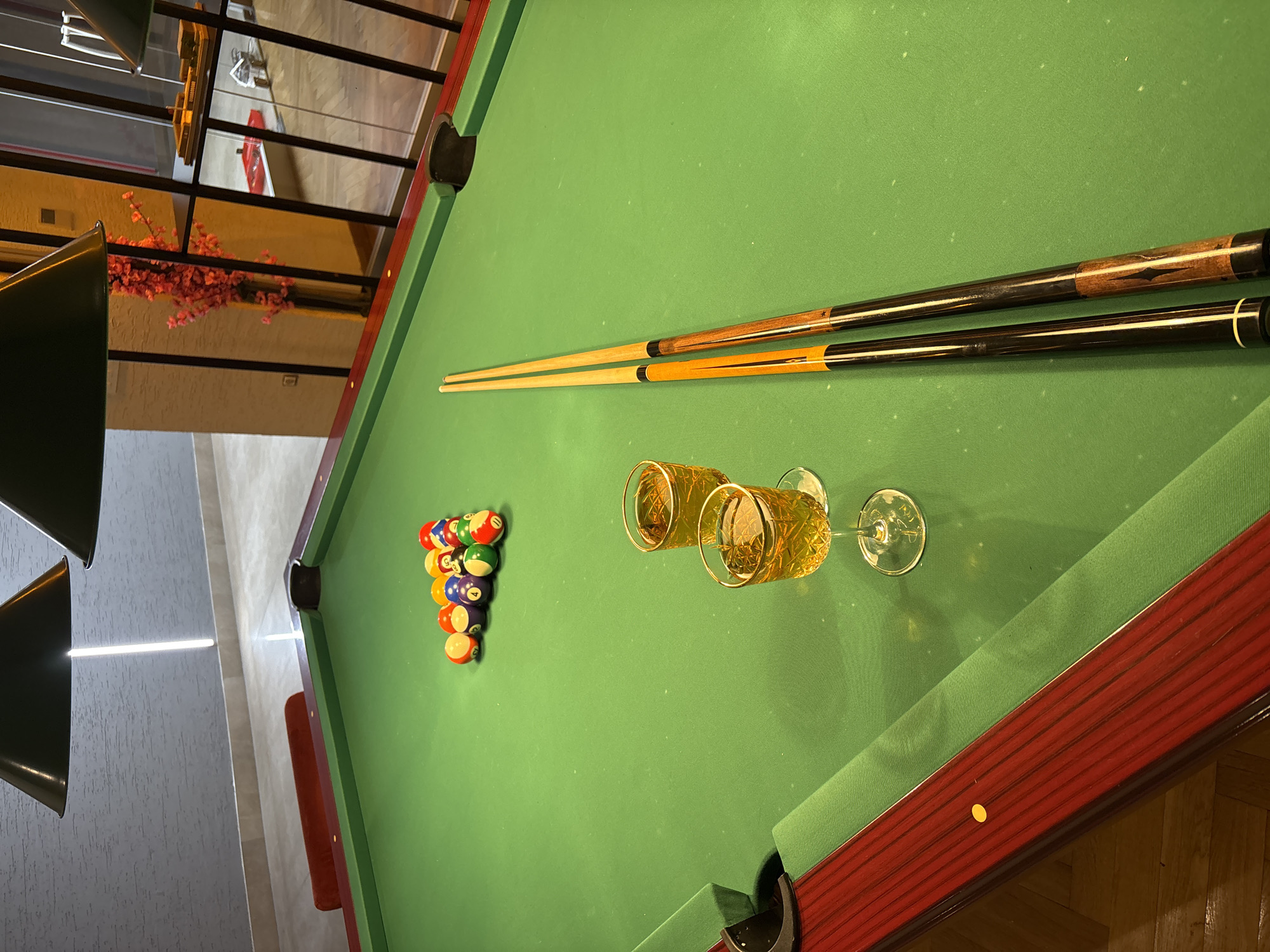 In Kâğıthane, Turkey, this image captures a close-up view of a green pool table, set for a game. Two billiard cue sticks lie on the felt, positioned parallel to each other. A rack of thirteen billiard balls, including solids and stripes, is arranged at the top of the table. Two stemmed glasses, filled with a golden liquid, sit on the green surface. The pool table is bordered by a dark red railing, with a yellow sticker visible on the left side. The background reveals a modern interior with textured walls and overhead lighting fixtures.