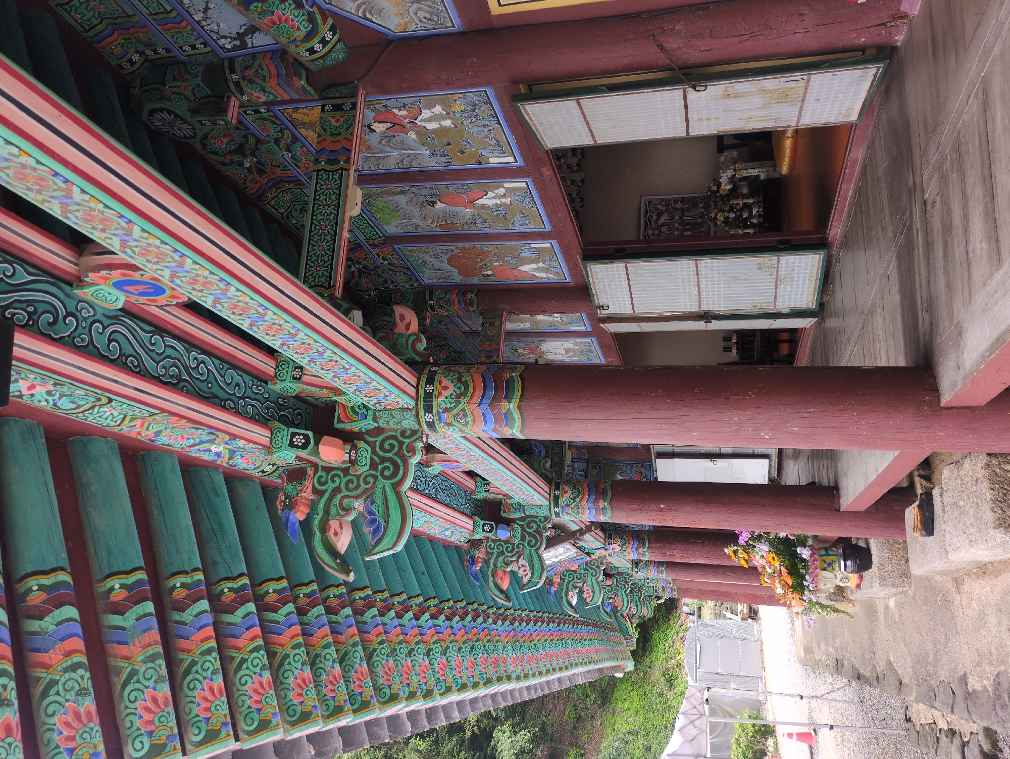 The image captures a section of the Gakwonsa Temple in Gongju-si, South Korea, focusing on its elaborately decorated eaves and support structure. The eaves feature rows of green, tiled roofs adorned with painted lotus flower motifs and layered, colorful designs. The supporting wooden structure is painted with intricate patterns in green, red, blue, and gold, with small depictions of figures on the ceiling panels. Heavy, unpainted wooden pillars support the structure, framing views into a building with traditional sliding doors and an altar. A flower arrangement sits near the base of one of the pillars. The background includes a paved area and greenery.