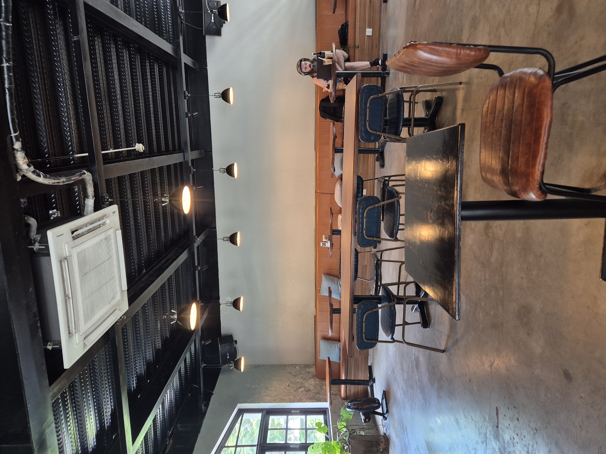 The image shows the interior of a modern cafe in Da Nang, Vietnam. The cafe features a high ceiling with exposed black metal beams and a white cassette air conditioning unit. Industrial-style pendant lights hang from the ceiling. The walls are a neutral light gray, with one section having a textured concrete finish.
The seating area includes a long, brown leather banquette that runs along the wall, with several light blue cushions placed on it. In front of the banquette are several wooden tables with black metal legs. In the foreground, a square black table with a thick edge is prominent, with a distressed brown leather chair with a black metal frame beside it. Further back, there are more wooden tables paired with dark blue chairs with black metal frames.
On the far right of the image, a man with a beard and headphones is seated at a table, looking at a laptop. He has a bottle of liquid and a glass next to him. Other items visible include a black backpack on the banquette behind him and a small bottle on a table near the man.
To the left, a large window with dark frames allows natural light into the space. A potted green plant is near the window, and a black oscillating fan stands on the concrete floor. The floor itself is a polished concrete surface. The overall ambiance is contemporary and somewhat industrial, typical of many modern cafes.