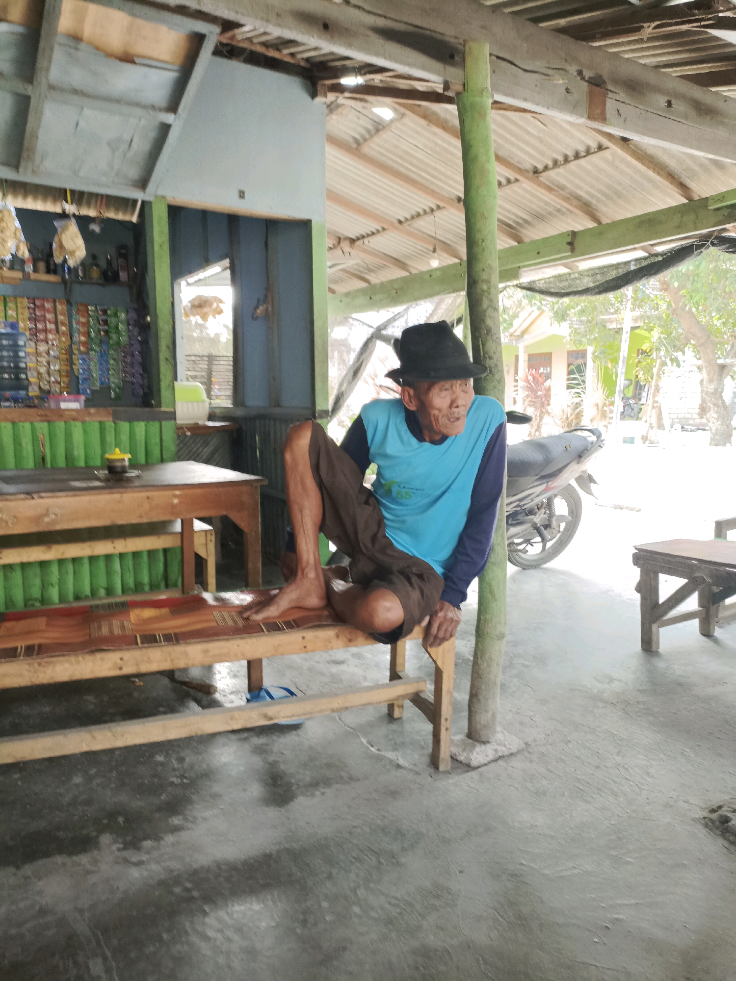 An elderly Indonesian man wearing a black hat and a blue long-sleeved shirt sits on a wooden bench in an open-air structure in Rancang Kencono, Indonesia. The structure appears to be a small shop or roadside stall, with shelves of packaged goods visible on the left and a table with a yellow object on it. A motorcycle is parked to the right of the man. The ground is concrete, and there are wooden supports for the roof. The man is looking to his right with his legs crossed.