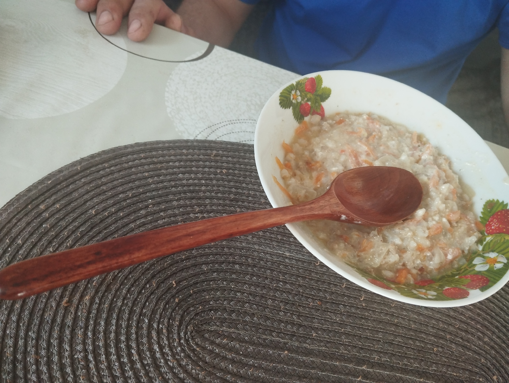 The scene depicts a partially eaten bowl of food with a wooden spoon resting on top of it. The food appears to be porridge with small pieces of orange vegetable, possibly carrots. The bowl is white with a strawberry pattern around the rim. The bowl rests on a dark brown woven placemat. A portion of a light-colored tabletop is visible in the background, as well as a person wearing a blue shirt. Part of another person's hand is also visible. The location is Krasnogorskoye, Russia.