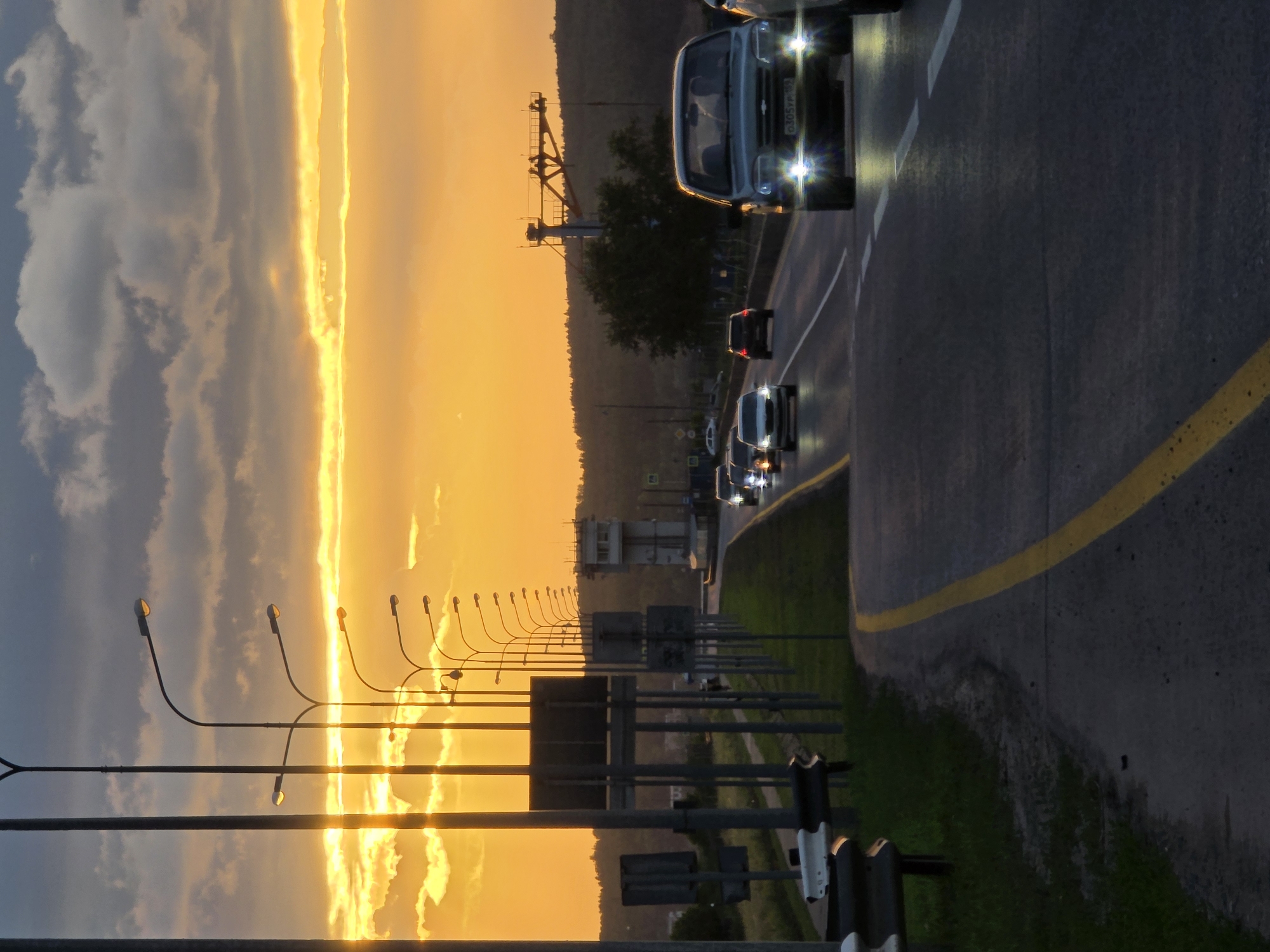 The image depicts a road scene at sunset, possibly on the outskirts of Chaikovsky, Russia, indicated by a Russian license plate on the foreground vehicle. The sky is a vibrant mix of orange and yellow hues, with scattered clouds, suggesting either sunrise or sunset. A highway stretches into the distance, with several cars traveling in the same direction. The road is marked with white and yellow lines. On the left side of the road, a series of streetlights line the shoulder, with some illuminated. A guardrail is visible in the foreground on the left. In the background, a control tower-like structure stands near the road, and a densely wooded hillside forms the horizon. The overall atmosphere is one of transition, with the fading light of day casting long shadows.