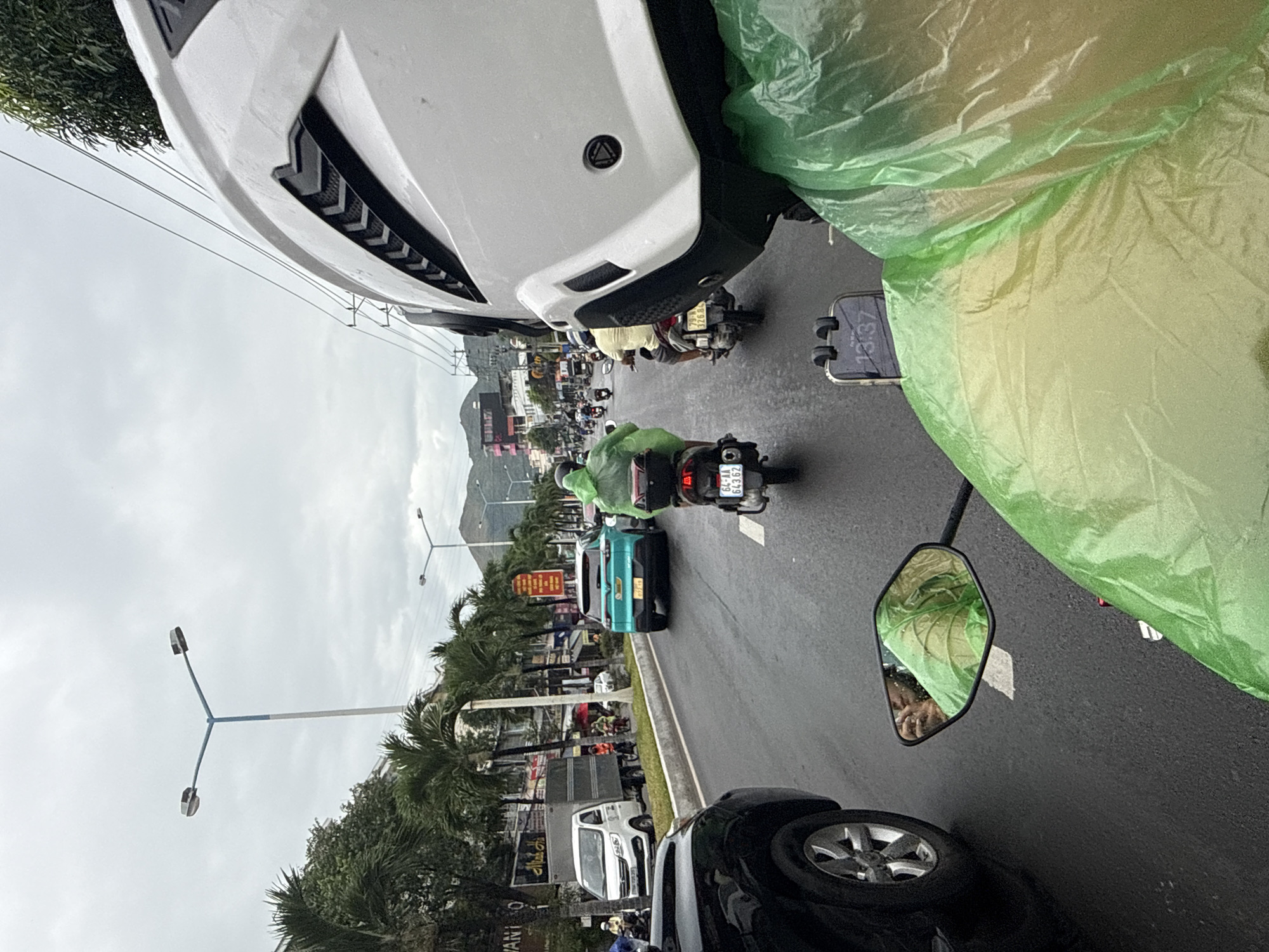 The image depicts a rainy day in Nha Trang, Vietnam, likely on a main road. The perspective is from a motorcycle rider, evidenced by the motorcycle's mirror reflecting the rider's face and hand, and the prominent white motorcycle helmet in the foreground. The rider is wearing a bright green rain poncho. The road is wet, indicating recent or ongoing rain, and is lined with palm trees and buildings. Several vehicles are visible, including a black SUV in the foreground, a white delivery truck, a teal-colored SUV, and numerous motorcycles. Most other riders are also wearing rain ponchos, many in green. Traffic appears to be moderate. The sky is overcast. The time "13:37" is visible on a phone mounted on the handlebars.