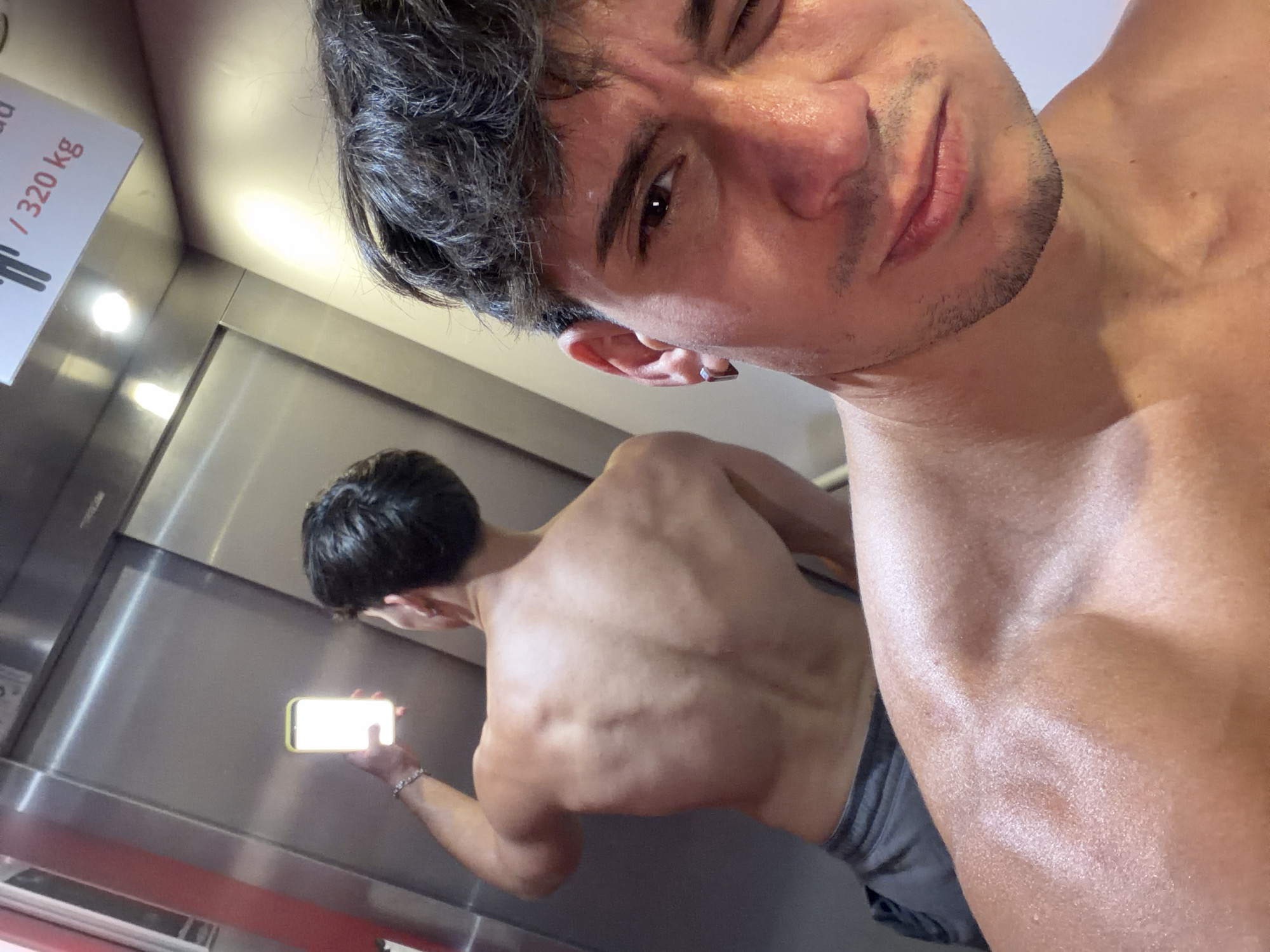 A young man with dark curly hair takes a selfie in an elevator, holding his phone in his left hand. He is shirtless, showing his back and shoulders, and wearing grey shorts. A mirror reflects his back, revealing well-defined muscles. The elevator has stainless steel walls and a sign indicating a maximum load of 320 kg. The man has a square earring in his left ear and appears to be sweating. The scene is brightly lit. The location is not specified as Sofia, Bulgaria.