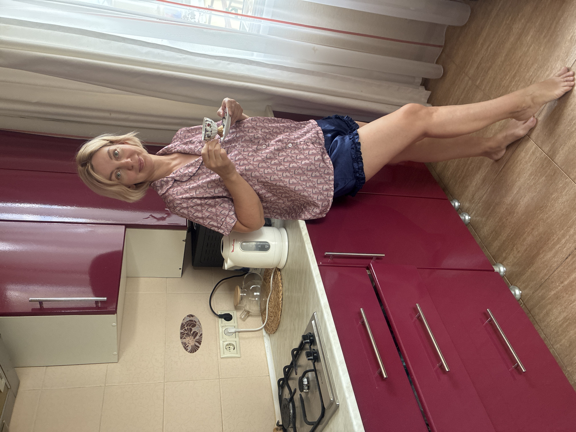 A woman stands in a kitchen, holding a teacup and saucer, near a window with white curtains. She is wearing a pink patterned pajama top and blue satin shorts. To her left is a counter with a gas stove, a glass teapot, and a white electric kettle. The kitchen features dark pink cabinets and cream-colored tiled walls. The flooring is light brown wood. The scene is set in a kitchen in Sochi, Russia.