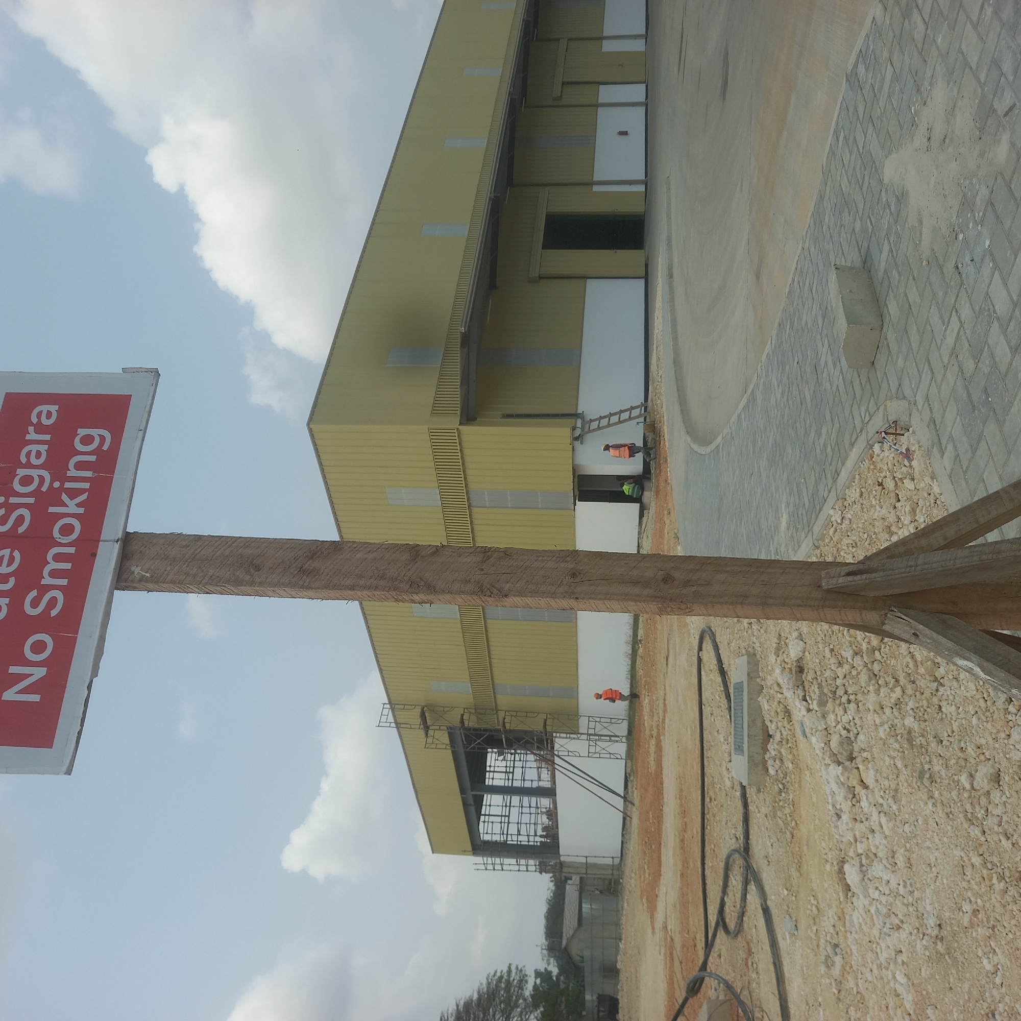In Bububu, Tanzania, a construction site is captured under a cloudy sky. A large, yellow industrial building, partially enclosed by scaffolding, dominates the scene. The building features white paneling at its base and a beige roofline. To the left, a smaller, grey metal structure is visible. Several construction workers in orange vests are present. One is near the yellow building, and two others are near the entrance. A wooden post with a "No Smoking" sign stands in the foreground, partially obscuring the view. The ground is a mixture of dirt, gravel, and a paved walkway with interlocking concrete blocks. Construction materials and equipment, including black hoses and a concrete electrical box, are scattered on the ground.