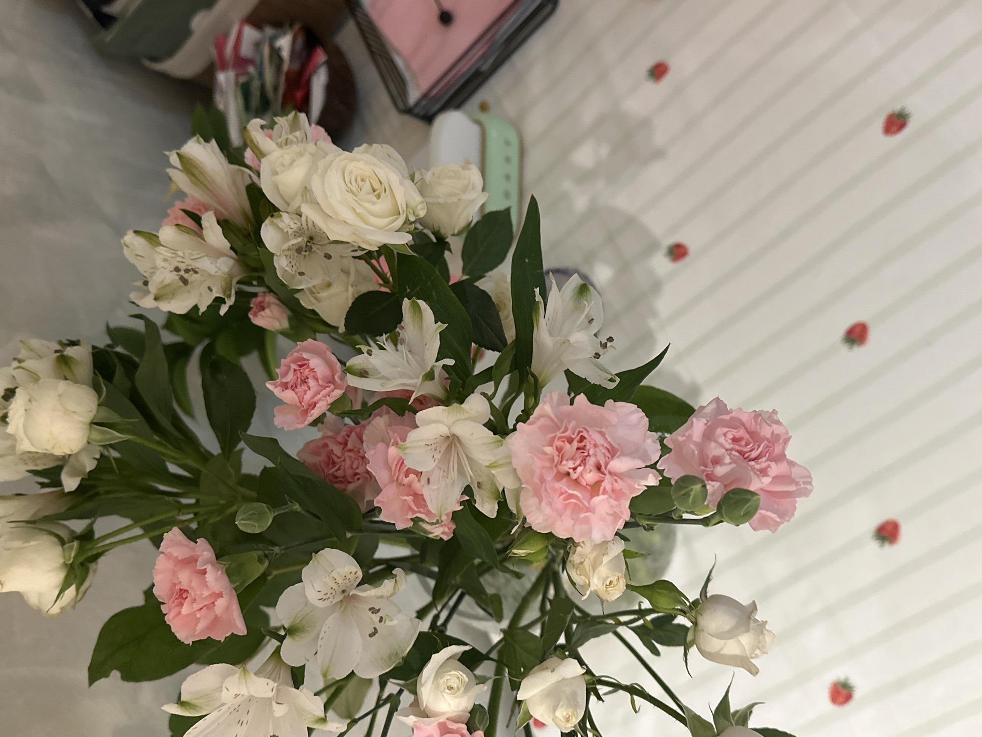 A high-angle, eye-level shot presents a floral arrangement featuring a variety of blooms in shades of pink and white. The bouquet consists of pink carnations, white roses, and white alstroemeria. The flowers are interspersed with green foliage. The backdrop is a white surface with subtle vertical stripes and small red strawberry patterns scattered across it. The lighting is diffused, casting soft shadows. In the upper-left corner, a glimpse of a blurred object suggests a cluttered background. The image was not taken in Šušanj, Montenegro, or it cannot be determined to be taken there based on the given information.