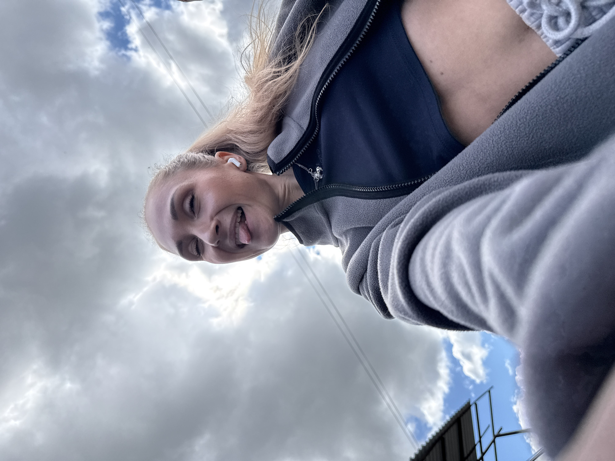 An upward-angled selfie captures a young woman outdoors in Chaikovsky, Russia, on a day with mixed cloudy and clear skies. The woman, with blonde hair pulled back, is visible from the chest up, positioned towards the left of the frame. She is wearing a grey zippered fleece jacket over a dark blue top. A white wireless earbud is in her right ear. She has a playful expression, smiling and sticking her tongue out towards the camera. A small, shiny pendant on a necklace is visible near the zipper of her jacket.
The background is dominated by a dynamic sky, featuring a mix of dark grey, overcast clouds and brighter white clouds, with patches of clear blue sky visible. Thin utility lines or power lines run diagonally across the sky from the upper right. In the bottom right corner, a portion of a metal-framed structure, possibly a building roof or billboard, is seen against a patch of blue sky. The overall scene conveys a casual and cheerful mood, with the low-angle perspective providing a unique view of the subject against the sky. No visible text is present in the image.