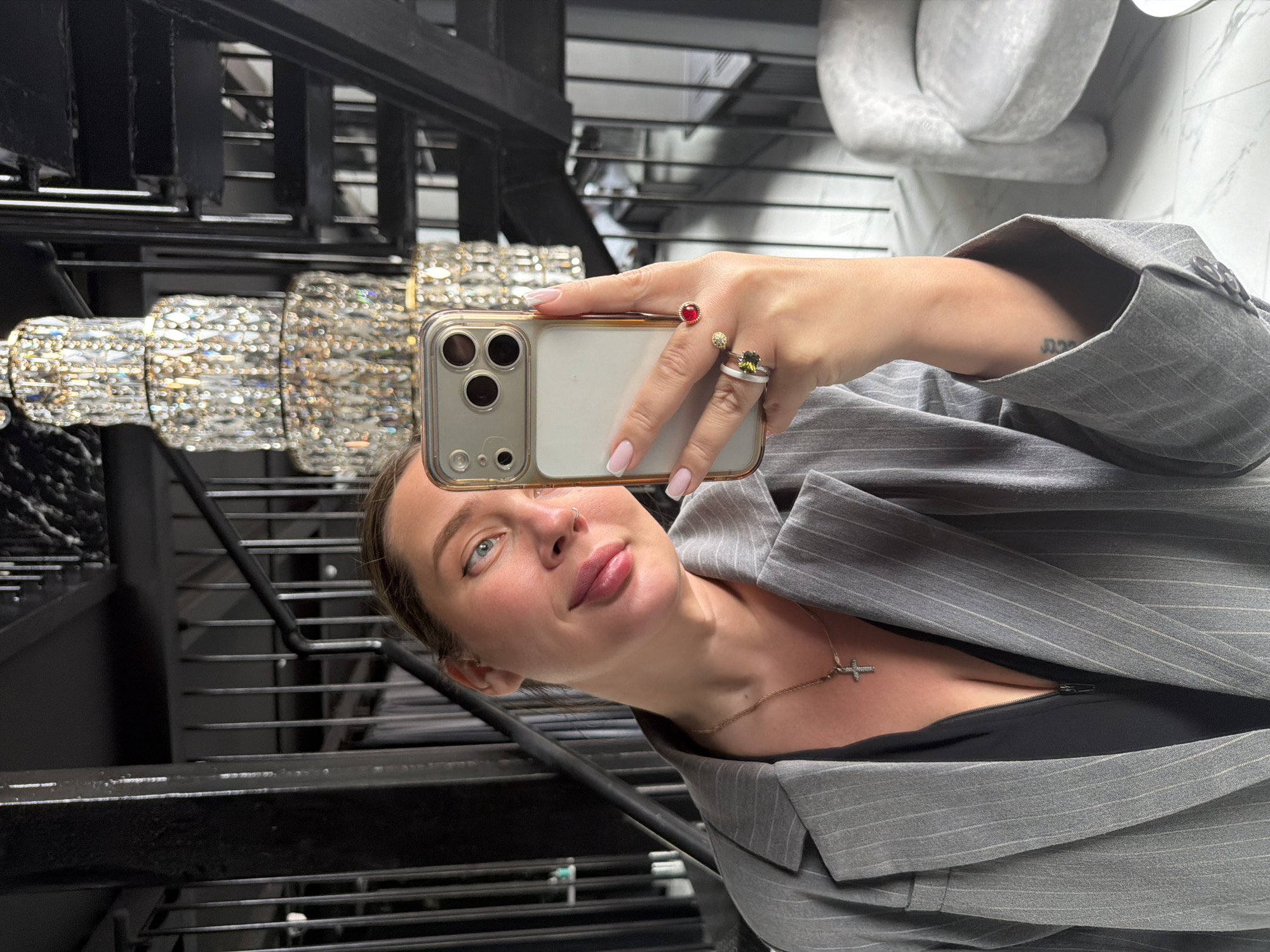 A woman in a gray pinstripe blazer is taking a selfie with an iPhone. She is wearing rings on her left hand and a gold necklace with a cross pendant. The background includes a crystal chandelier, black metal railings, and a white marble wall. The location is possibly in Podolsk, Russia, given the prompt's mention of the city.
