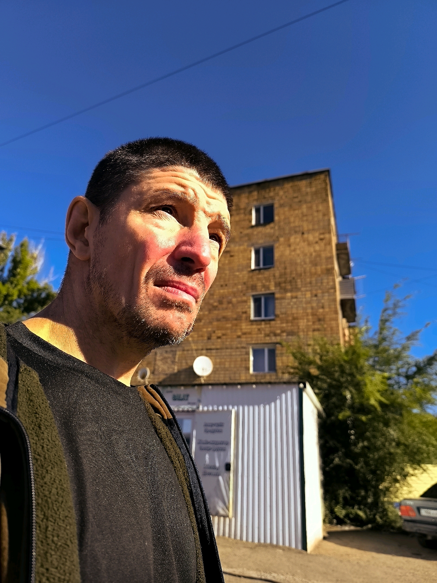 A middle-aged man with short dark hair and stubble is looking up and to the right, with sunlight illuminating the right side of his face. He is wearing a dark jacket over a black garment. In the background, a multi-story brick building with several windows stands under a clear blue sky. To the left of the building is a smaller white structure with vertical siding, and a satellite dish is visible on the side of the brick building. Lush green trees are present to the right of the man, and a portion of a car is visible on the far right. The scene appears to be outdoors in an urban or suburban setting.