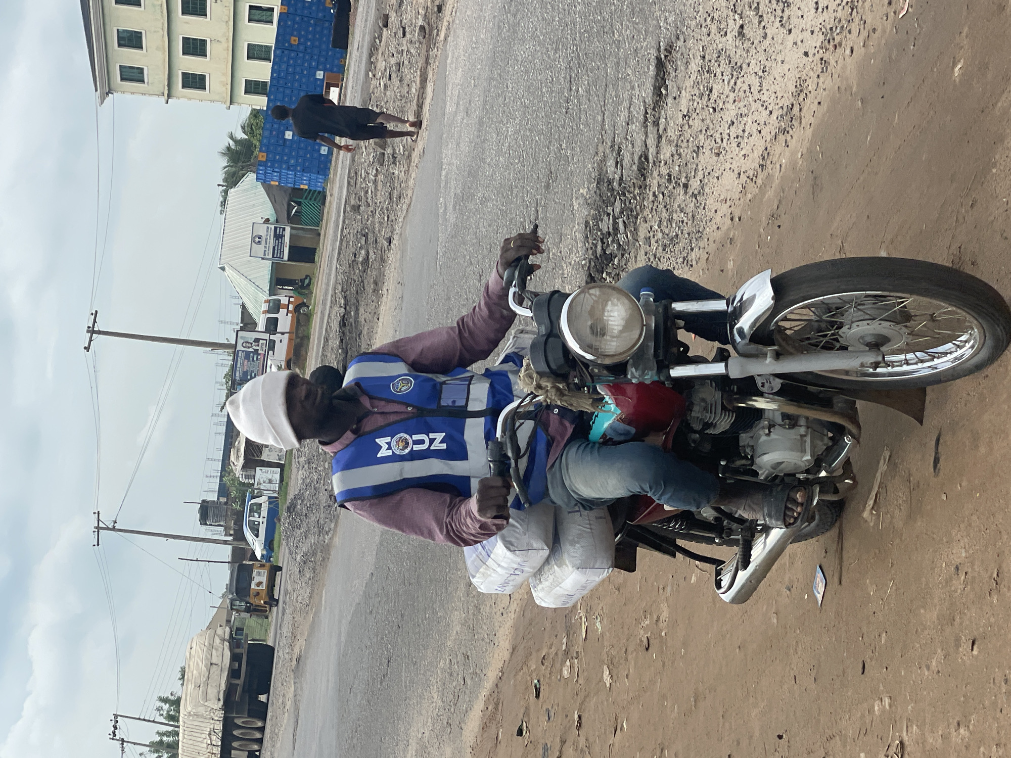 A man wearing a white beanie, a pink collared shirt, blue jeans, and a blue high-visibility vest with reflective stripes is riding a red motorcycle on a dusty road in Lokoja, Nigeria. He is carrying two large white bags, possibly cement, stacked behind him. The motorcycle has a chrome headlight and front fender. In the background, a large white truck with a covered load is visible on the left, along with a yellow tricycle and a white and blue van. Further back, there are utility poles, power lines, buildings, and stacks of blue containers. The road surface is uneven and appears to be under construction or in disrepair. Another person in a dark shirt and shorts is walking away from the camera on the right side of the image. The sky is overcast.