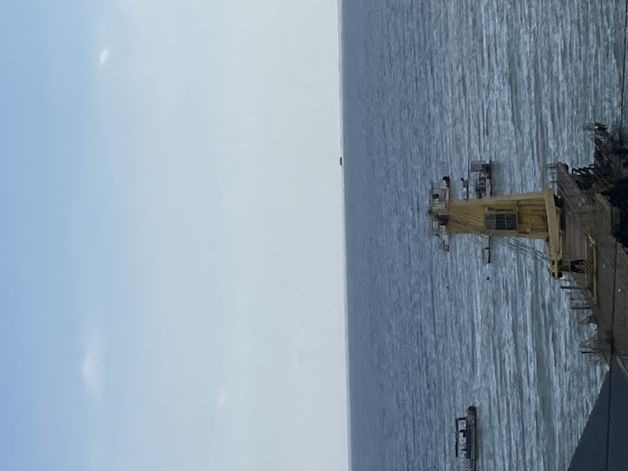 The image depicts a serene, overcast day at sea, likely in the vicinity of Sochi, Russia, given the presence of maritime structures typical of a port or coastal area. The scene is dominated by the vast expanse of the sea and sky.
In the foreground, partially visible at the bottom and right, is a section of a large, derelict yellow structure, possibly part of a disused industrial facility or a ship's superstructure, showing signs of decay with exposed metal and scaffolding. This structure occupies the lower right portion of the frame, with a dark roofline extending from the bottom left.
The middle ground is filled with the blue-gray, slightly rippling waters of the sea. Several small boats are scattered across the water. To the left, a flat-topped boat with a dark canopy is visible. Further out, another similar boat is visible. In the center, a tall, yellow structure, likely a crane or part of a pier, rises from the water. Two smaller boats are positioned near this structure.
In the distance, a thin, dark line on the horizon suggests a landmass or another vessel. The sky above is a pale blue, streaked with wispy white clouds, creating a diffused light over the entire scene. The overall impression is one of stillness and industrial decay against the backdrop of the open sea. There are no visible people in the image.