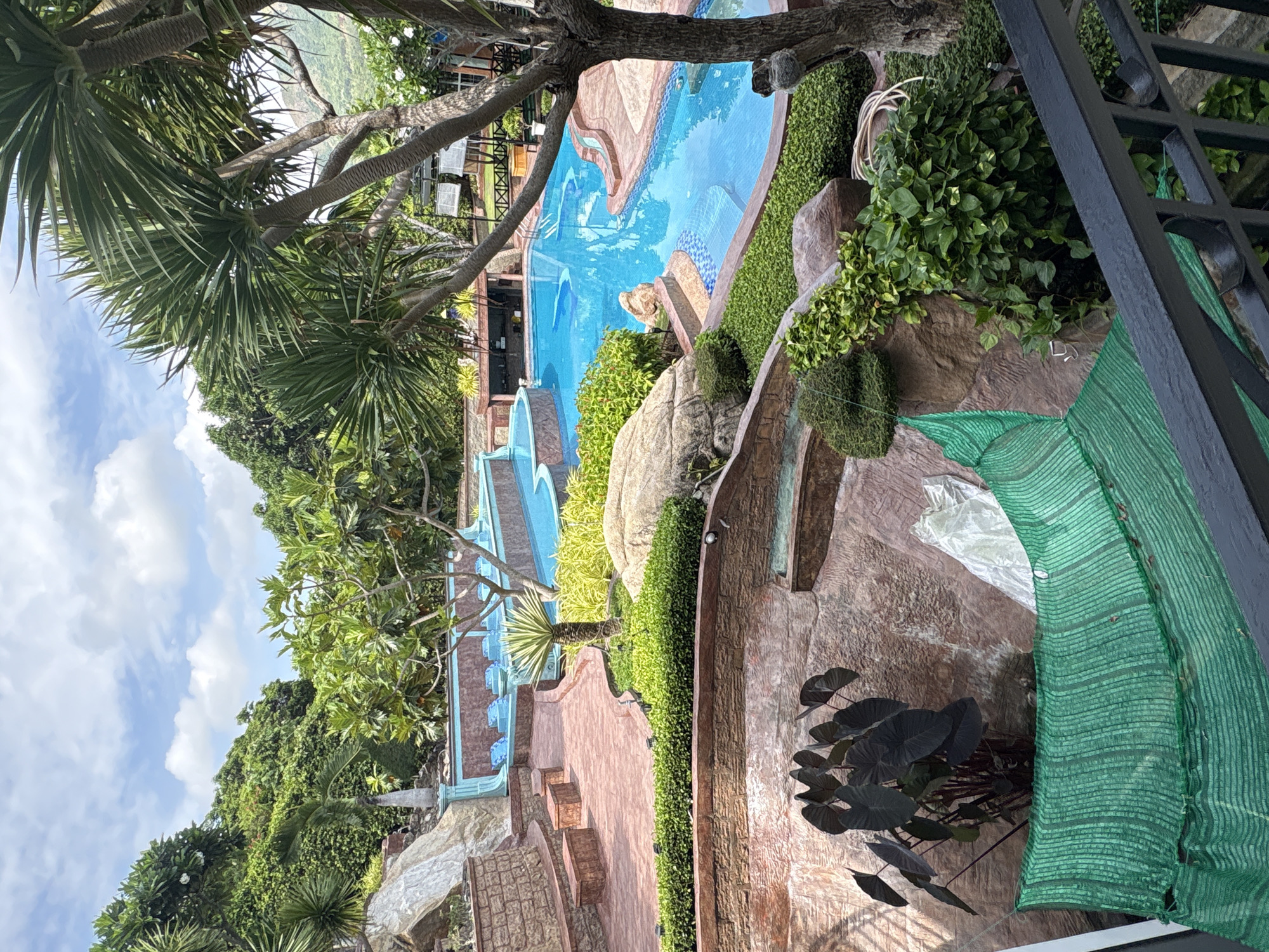 The image depicts an outdoor scene, possibly a resort or private residence, located in Bac Nha Trang Ward, Vietnam. The scene is dominated by a multi-tiered swimming pool complex with bright blue water. Lush greenery surrounds the pools, including various trees and shrubs. A tree branch partially obstructs the view in the upper portion of the frame. In the foreground, a set of metal railings or a fence is visible, along with a large green netting or covering. The surrounding landscape features hills covered in dense vegetation. The sky is partially visible, with scattered clouds.