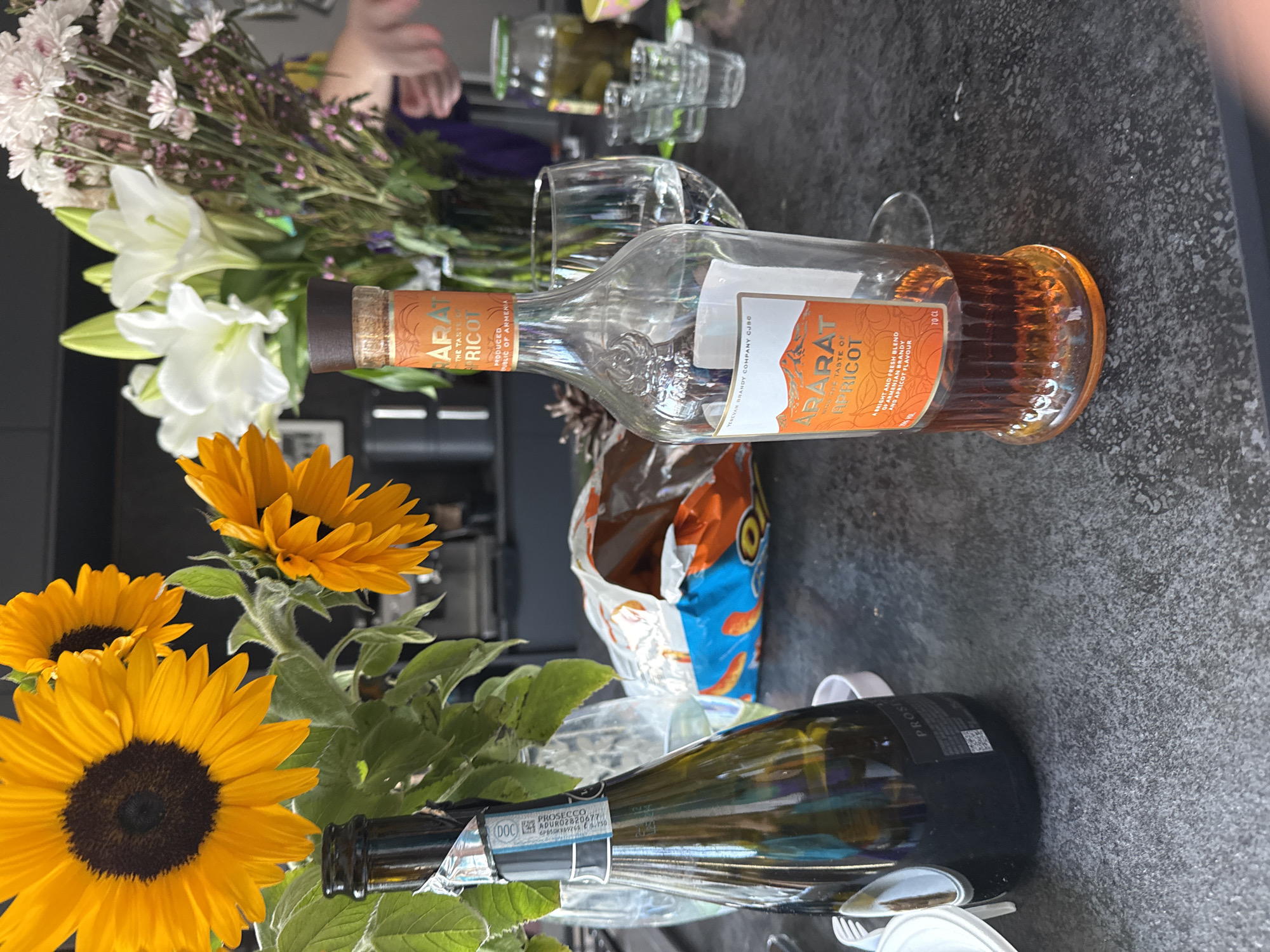 The scene is a tabletop with a variety of objects and natural elements. A bouquet of sunflowers and other flowers are prominent on the left side of the frame. A mostly empty bottle of ARARAT Apricot brandy is positioned with its label visible. Next to it, there is a clear drinking glass and an open bag of snacks. In the background, another bouquet of white lilies and smaller purple flowers can be seen. The scene is indoors, likely in a home setting. The city of Tel Aviv and the country of Israel are not evident in the image.