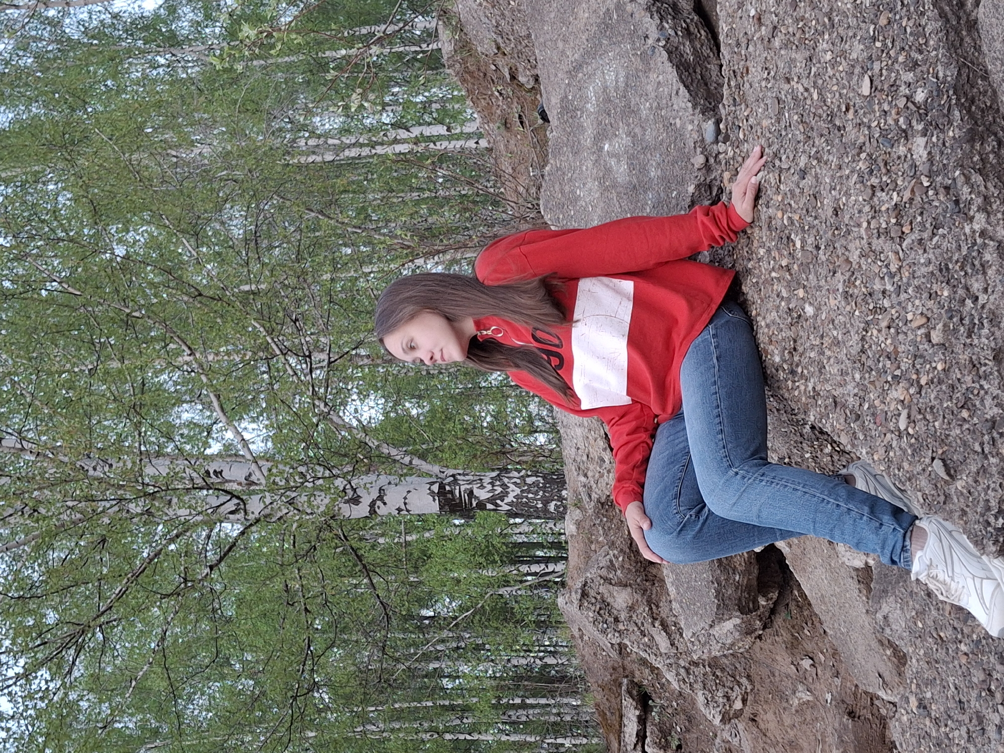 A young woman is sitting on a rocky outcropping near a birch forest in Chaikovsky, Russia. She is wearing a red long-sleeved sweatshirt with a white stripe across the chest, blue jeans, and white sneakers. She is leaning against a large boulder with her hand resting on its surface. The background consists of a dense forest of birch trees with green leaves. The scene is outdoors during daylight.