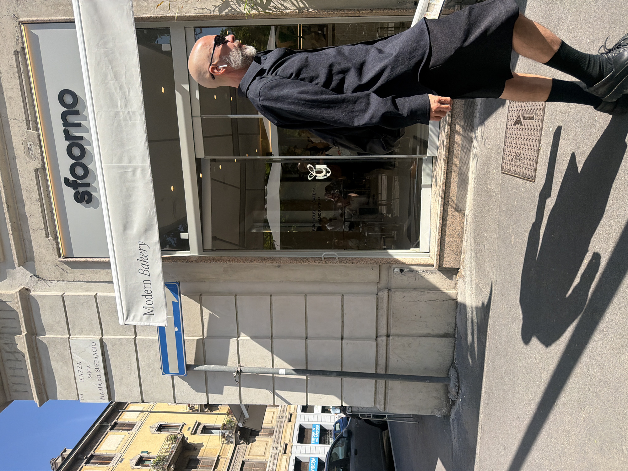 A man stands outside Sfoorno Modern Bakery in Milan, Italy, at Piazza Santa Maria Del Suffragio. He wears a black button-down shirt, dark skirt, black socks, and black shoes. Sunglasses rest on his head, and a white earbud is in his ear. Sunlight casts shadows on the sidewalk beside the building. A blue directional sign is positioned beneath the bakery's awning. Across the street, buildings can be seen under a bright sky. A black car is parked along the street.