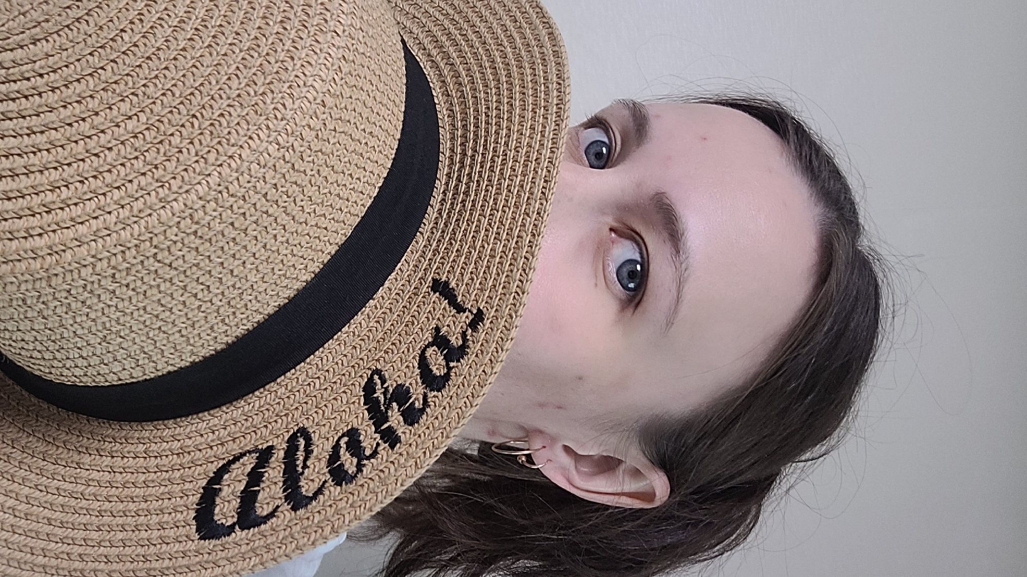 A woman is partially obscured by a straw hat, her eyes visible above it. The hat has a black ribbon band and appears to have embroidered text that reads "Beach" in black lettering. She has fair skin and dark brown hair pulled back into a ponytail. She is wearing small hoop earrings. The background is a plain, light-colored wall. The image is a close-up portrait, with the woman positioned in the center, looking towards the camera.