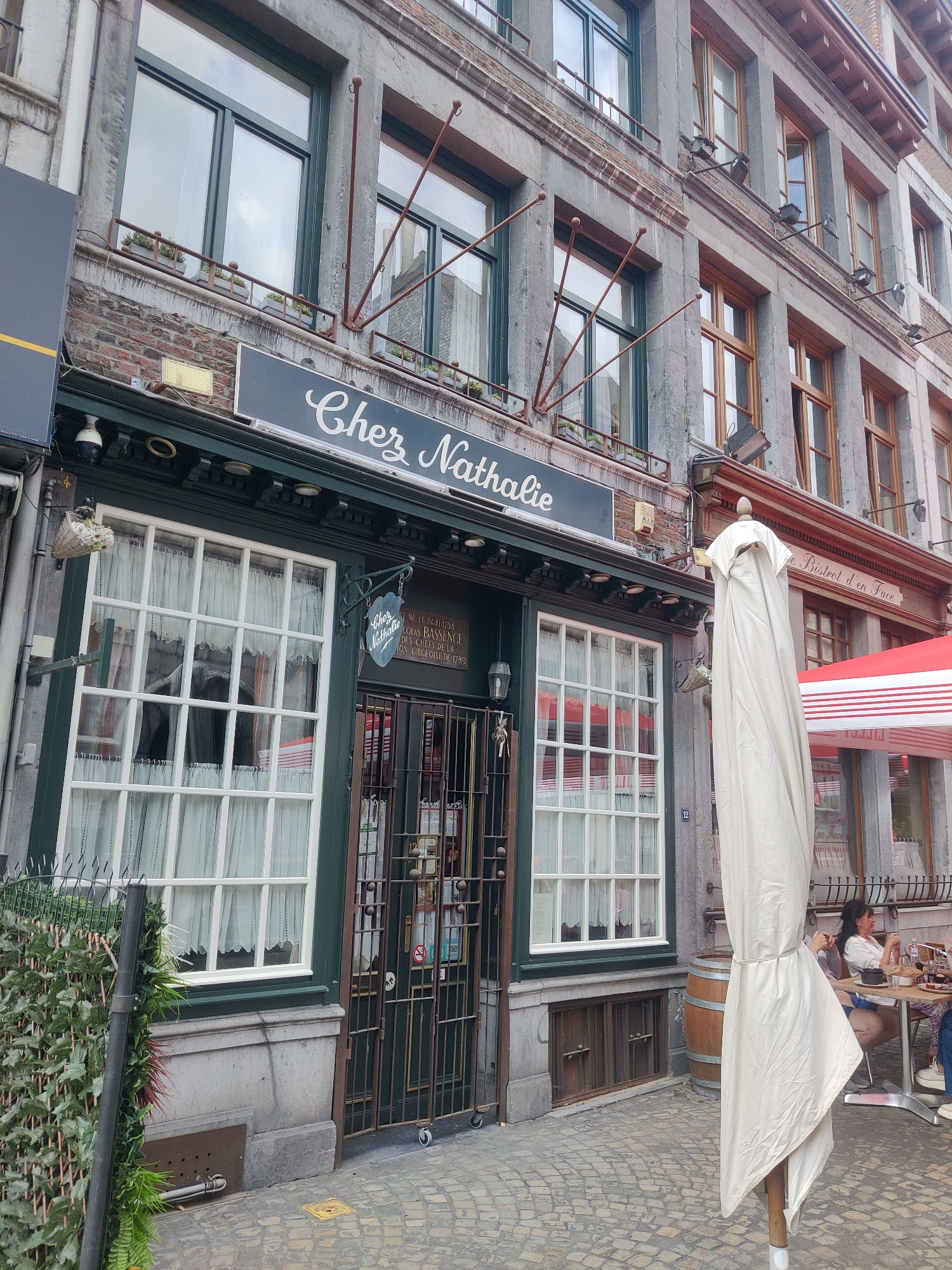 The image shows the exterior of a restaurant named "Chez Nathalie" located on the Rue de l'Étuve in Liège, Belgium. The restaurant features a green facade with large, multi-paned windows covered by white curtains. A dark blue sign with white script reads "Chez Nathalie". Next to the entrance, a decorative sign in a shield shape also reads "Chez Nathalie" and contains text in French below it, including a date and reference to Liège. The entrance is secured by a metal gate. To the right of the restaurant, an umbrella is closed and covered with a white fabric. A table with two people is visible in the background, suggesting outdoor seating. The building's facade is made of grey stone and brick, with visible architectural details like window frames and decorative metalwork. The street is paved with cobblestones.