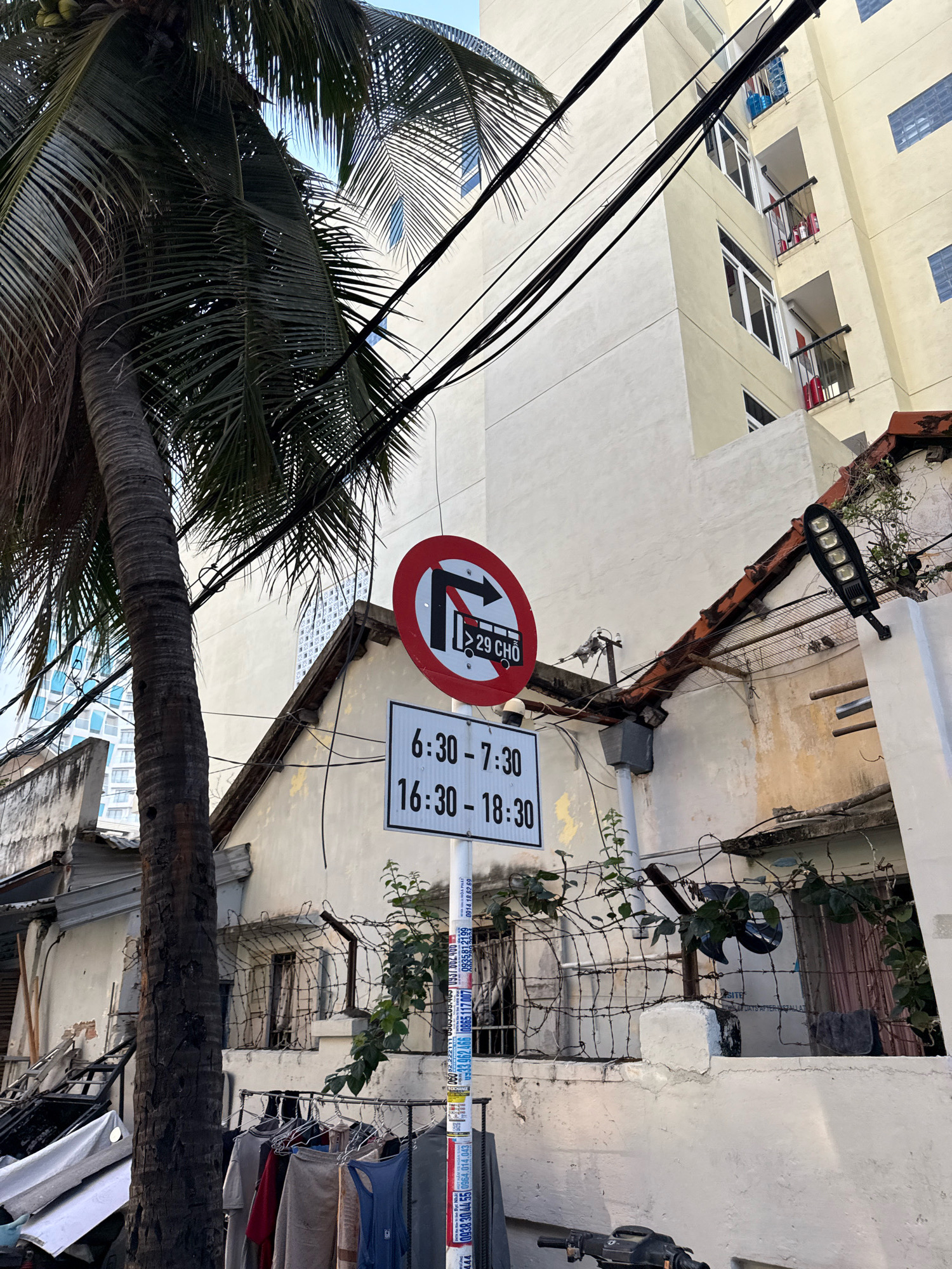 This image depicts a street scene in Nha Trang, Vietnam, featuring a traffic sign indicating restrictions for buses. The sign, a circular red-bordered prohibition sign, displays a silhouette of a bus with an arrow pointing left, suggesting that buses are not allowed to turn left at this location. Below this, a rectangular sign with white background and black text specifies times: "6:30 - 7:30" and "16:30 - 18:30", likely indicating when this restriction is in effect.
In the foreground, a portion of a palm tree is visible, its fronds extending towards the top of the frame. The background shows a light-colored multi-story building with balconies and windows. A tangle of electrical wires crisscrosses the scene, obscuring parts of the sky and the buildings. The environment appears somewhat cluttered, with a barbed-wire fence and some hanging clothing visible near the bottom of the frame, suggesting a residential or mixed-use area. The overall impression is that of an urban street environment in Vietnam, with typical elements like traffic signage, overhead wires, and local vegetation.