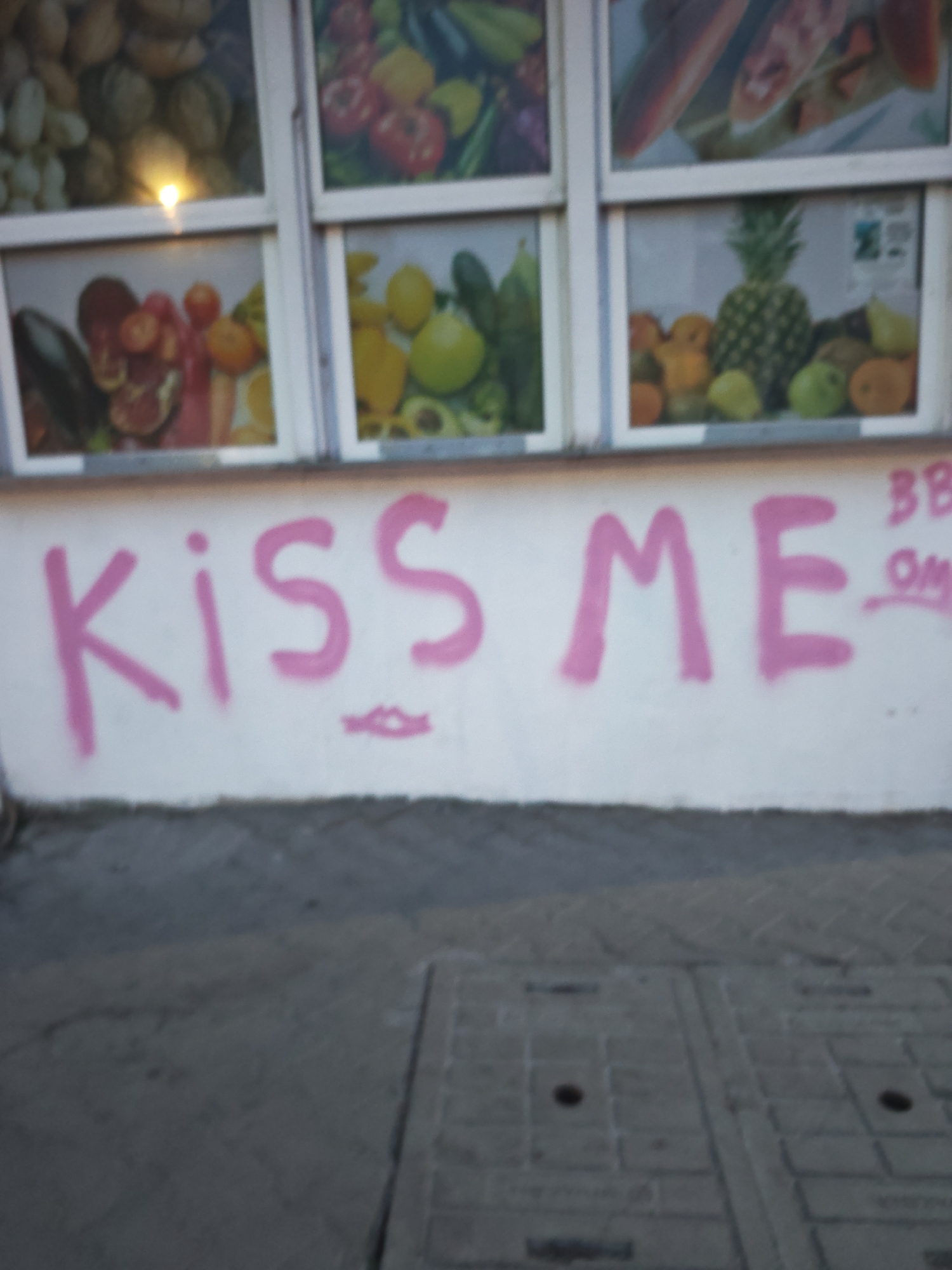 A white wall with pink graffiti spelling "KISS ME" is in the foreground of the image. The graffiti is in a handwritten style with thick strokes. Below the word "KISS," a pink drawing of lips is present. The wall is below a series of windows displaying images of fruits and vegetables. The windows are framed in white. The overall scene appears to be outdoors, possibly in front of a shop or food stand. The ground in front of the wall is a grey paved surface with a visible manhole cover.