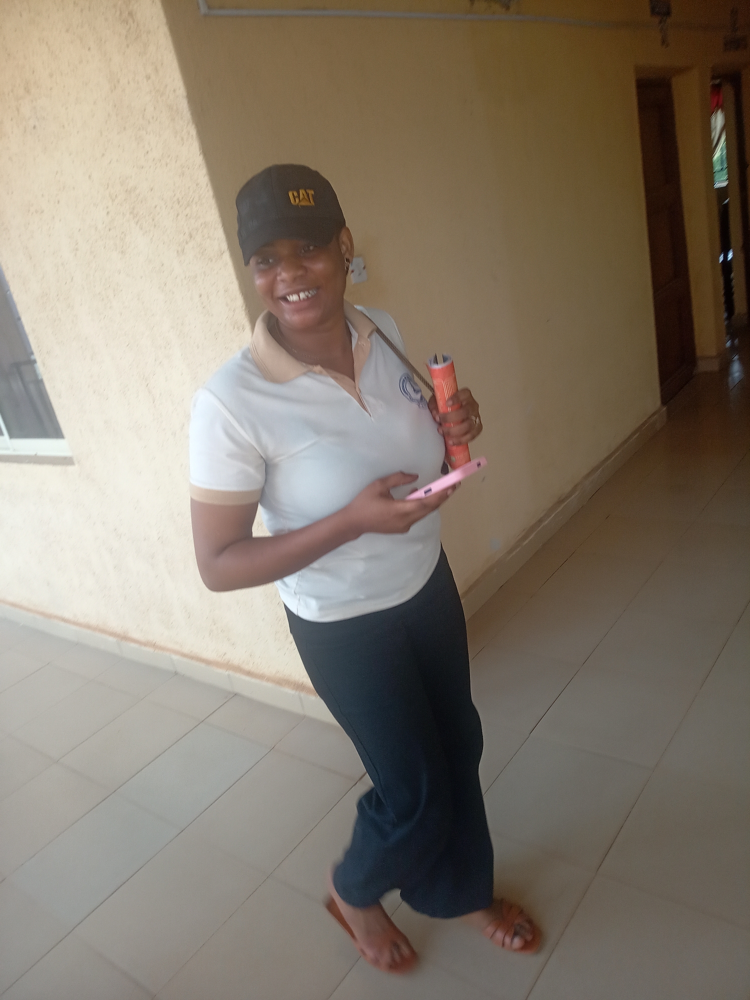 The image displays a woman smiling brightly in what appears to be an indoor corridor or covered walkway in Nkwelle, Nigeria. She has dark skin and is captured from the waist up, facing slightly towards the camera with a wide, toothy smile. She is wearing a black baseball cap with a yellow "CAT" logo on the front, a short-sleeved white polo shirt with beige accents on the collar and sleeve cuffs, and dark pants. Her feet are clad in light brown open-toed sandals.
In her left hand, she holds a pink smartphone, while her right hand holds an orange cylindrical object, possibly a rolled-up document or a container. A ring is visible on a finger of her right hand, and a strap, possibly from a bag, is seen over her right shoulder.
The background consists of light beige or yellowish-brown textured walls. A white-framed window is partially visible on the left, letting in natural light. The floor is covered with light-colored, rectangular tiles laid in a diagonal pattern. To the right, a darker opening with a wooden doorframe suggests an entrance to another room or hallway. The scene is well-lit, indicating it is daytime. The woman's happy and approachable demeanor is a notable detail.